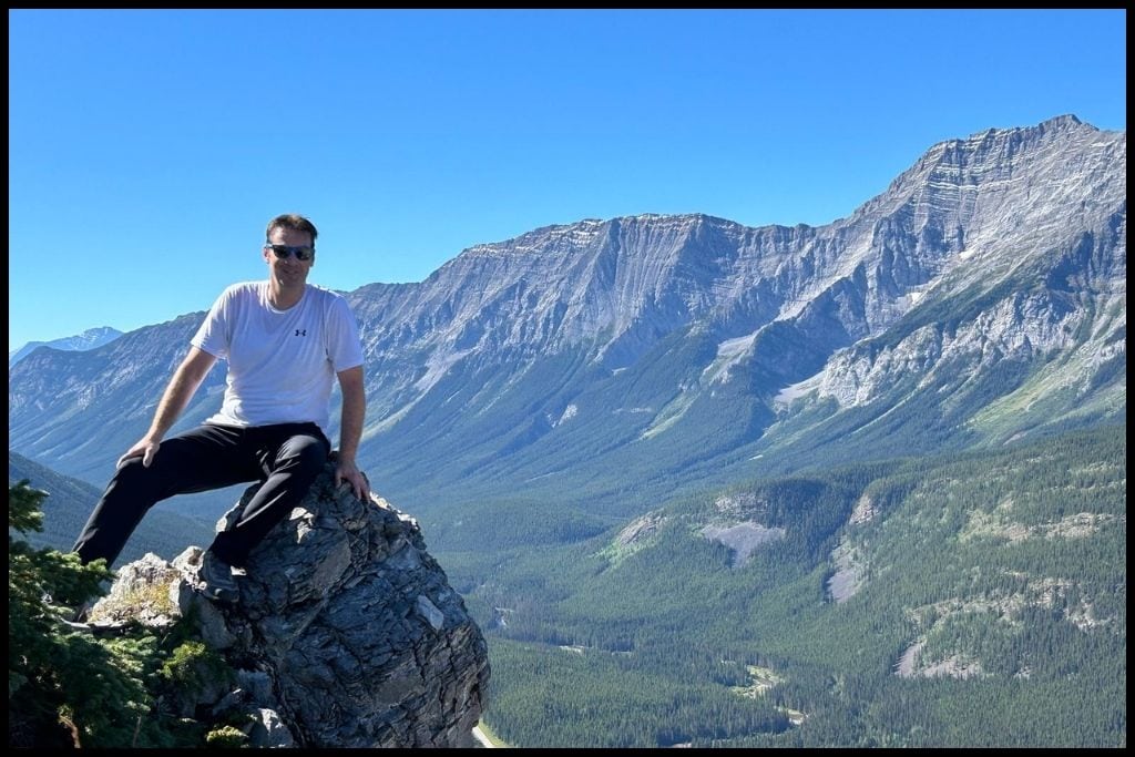 Man sitting on a rocky peak with panoramic views of Kananaskis mountains near Calgary, Alberta — showcasing outdoor recreation and hiking opportunities.
