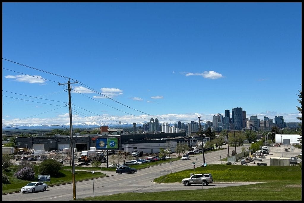 Clear view of downtown Calgary skyline on a sunny day, with modern high-rises set against the Rocky Mountains and blue skies, seen from a suburban vantage point.
