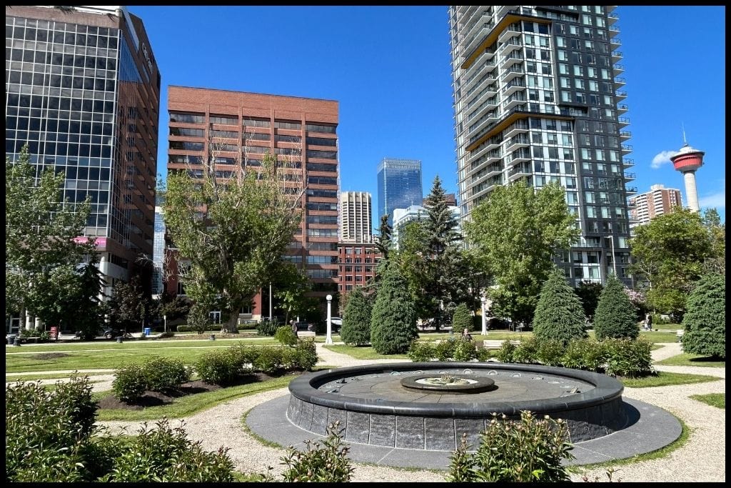 Scenic view of downtown Calgary featuring a central fountain, lush greenery, and modern high-rises, with the iconic Calgary Tower visible against a clear blue sky.