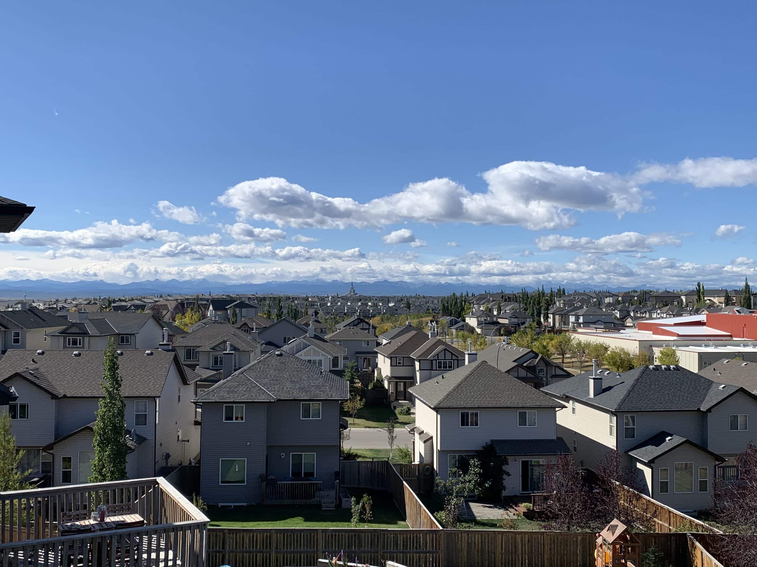 Scenic view of a suburban Calgary neighbourhood with detached homes, backyards, and a backdrop of the Rocky Mountains under a blue sky with scattered clouds.