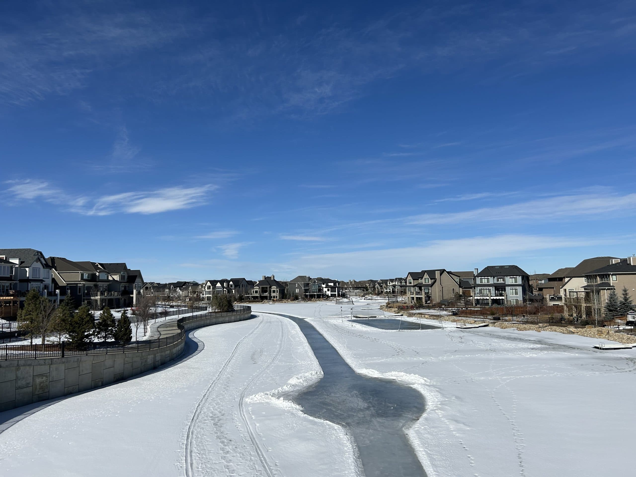 Winter view of Mahogany Calgary with a frozen lake and skating trail surrounded by modern homes under a bright blue sky.