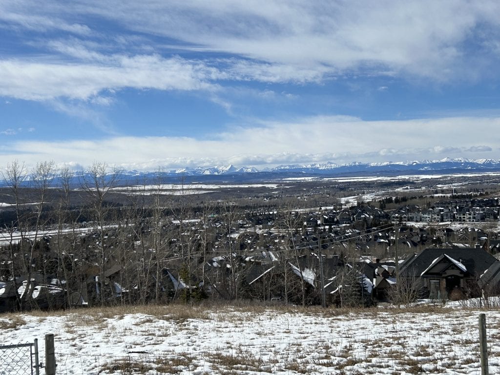 Winter view from Springbank Hill Calgary overlooking estate homes, snow-covered foothills, and the Rocky Mountains beneath a dramatic sky.
