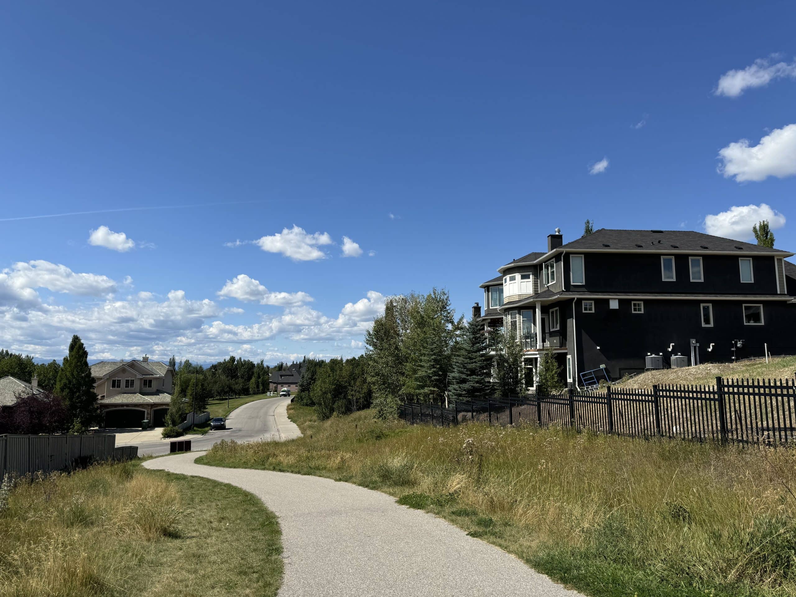 Paved pathway in Springbank Hill Calgary with upscale homes, mature trees, and blue skies in the background.