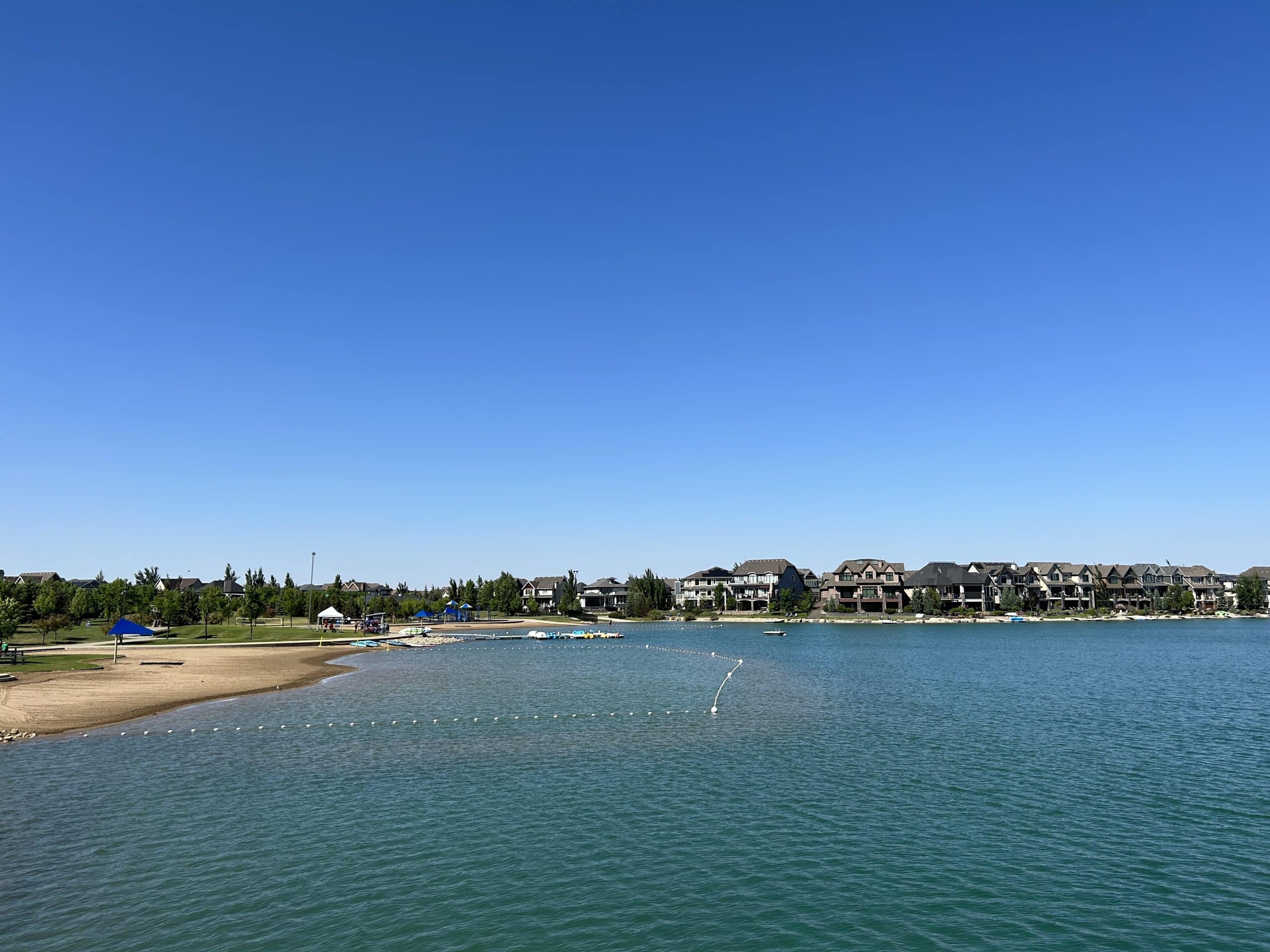 Mahogany Lake beach in Calgary featuring calm water, swimming area, paddleboat rentals, and lakeside homes under a clear summer sky.
