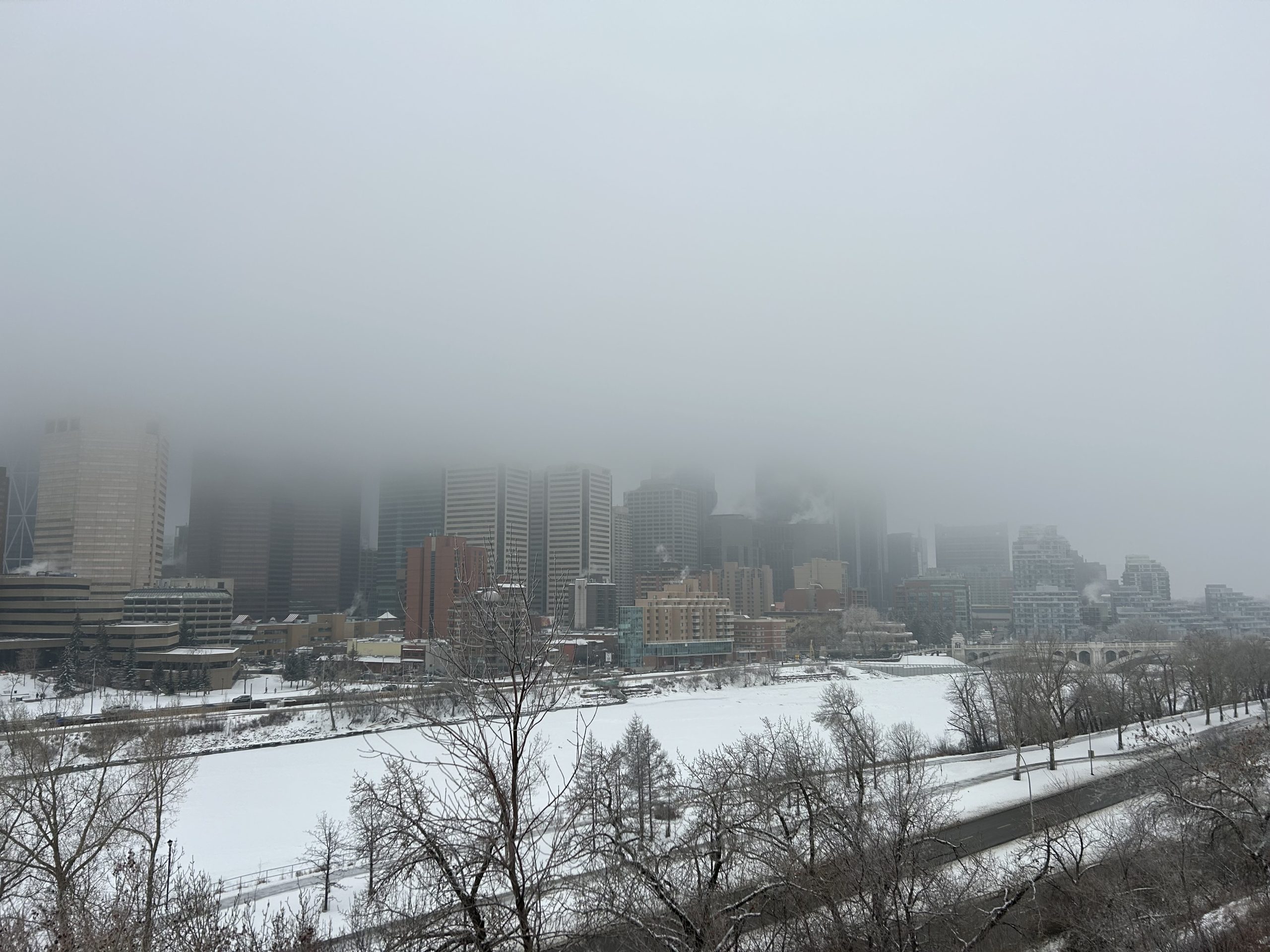 Fog covers downtown Calgary on a snowy winter day, with the Bow River frozen in the foreground. A striking view of Calgary’s skyline and harsh winter weather.