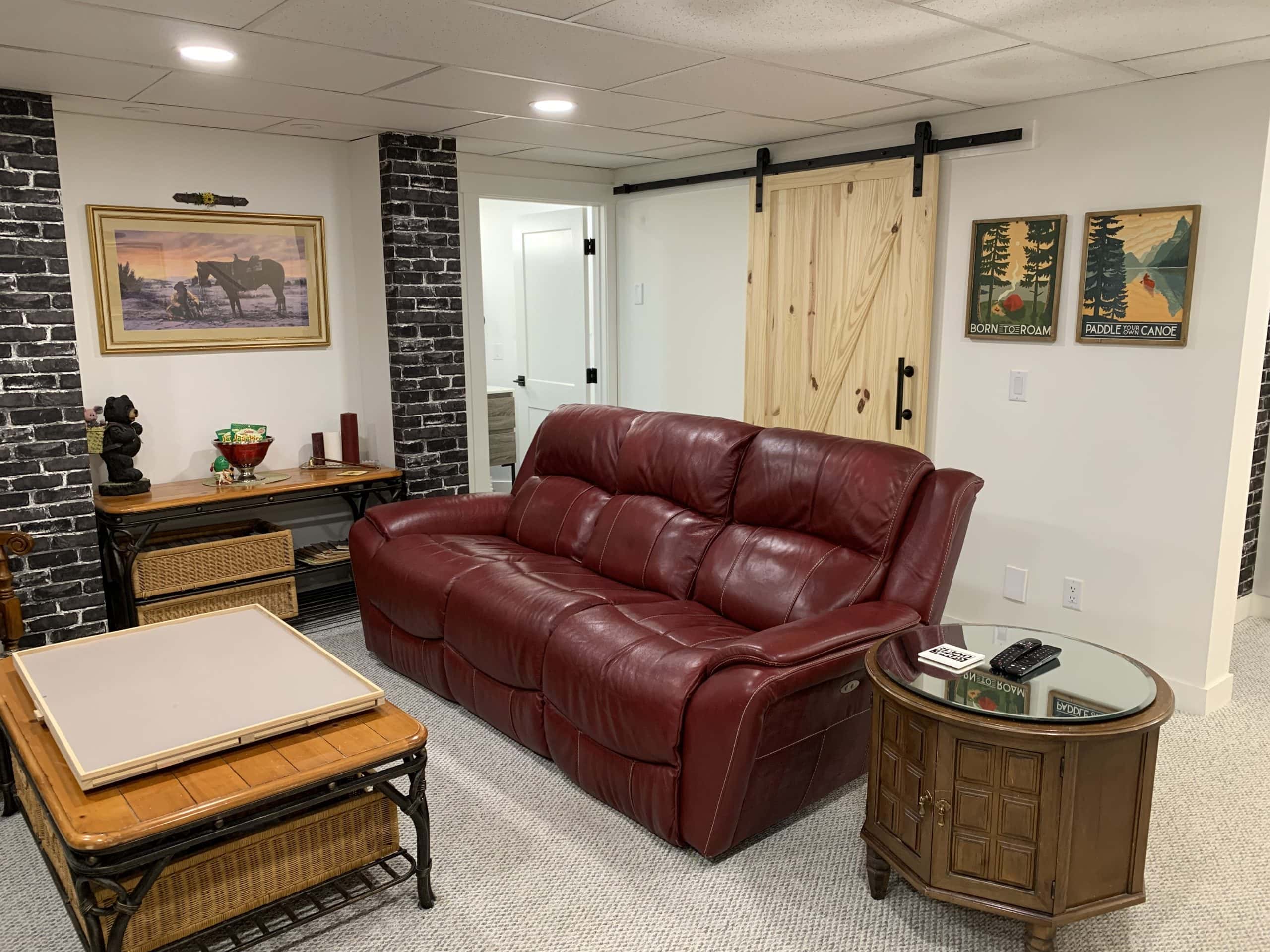 Cozy basement living room with a red leather sofa, rustic wood furniture, barn-style sliding door, and decorative artwork on the walls.