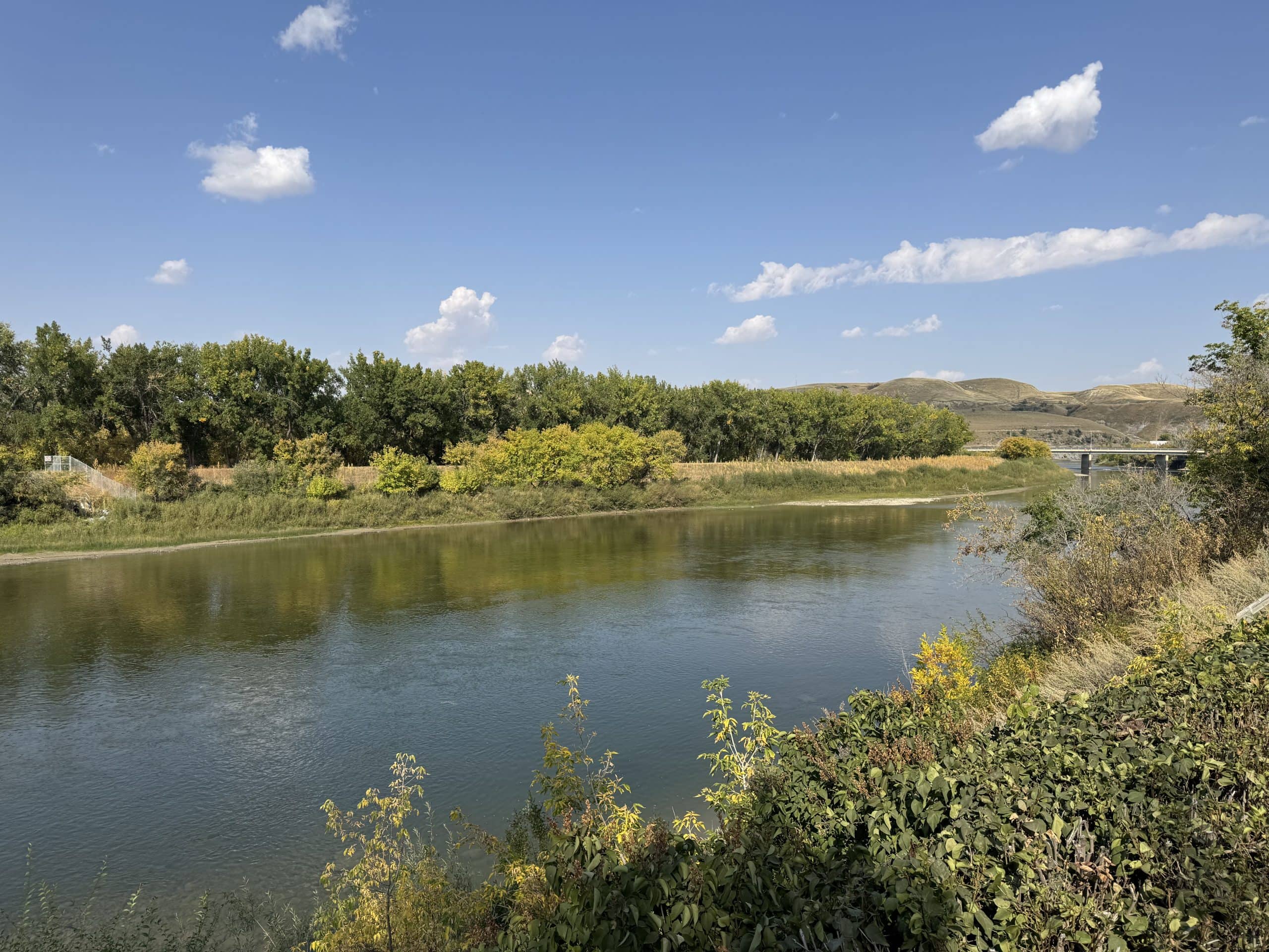 Scenic river view with clear blue water, lush green trees, and rolling hills under a bright sky with scattered clouds.