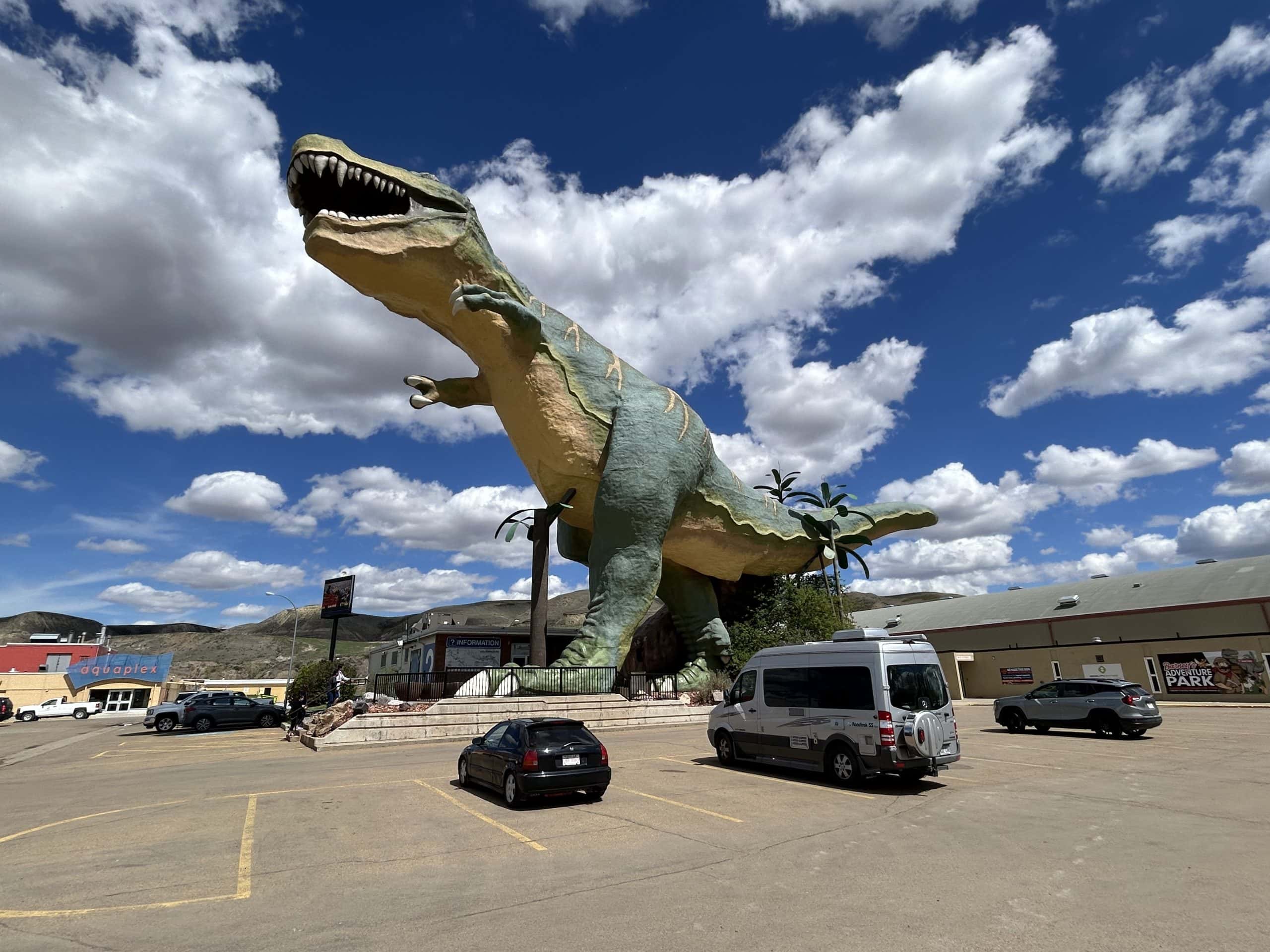Giant Tyrannosaurus rex statue towering over a parking lot in Drumheller, Alberta, set against a bright blue sky with scattered clouds.