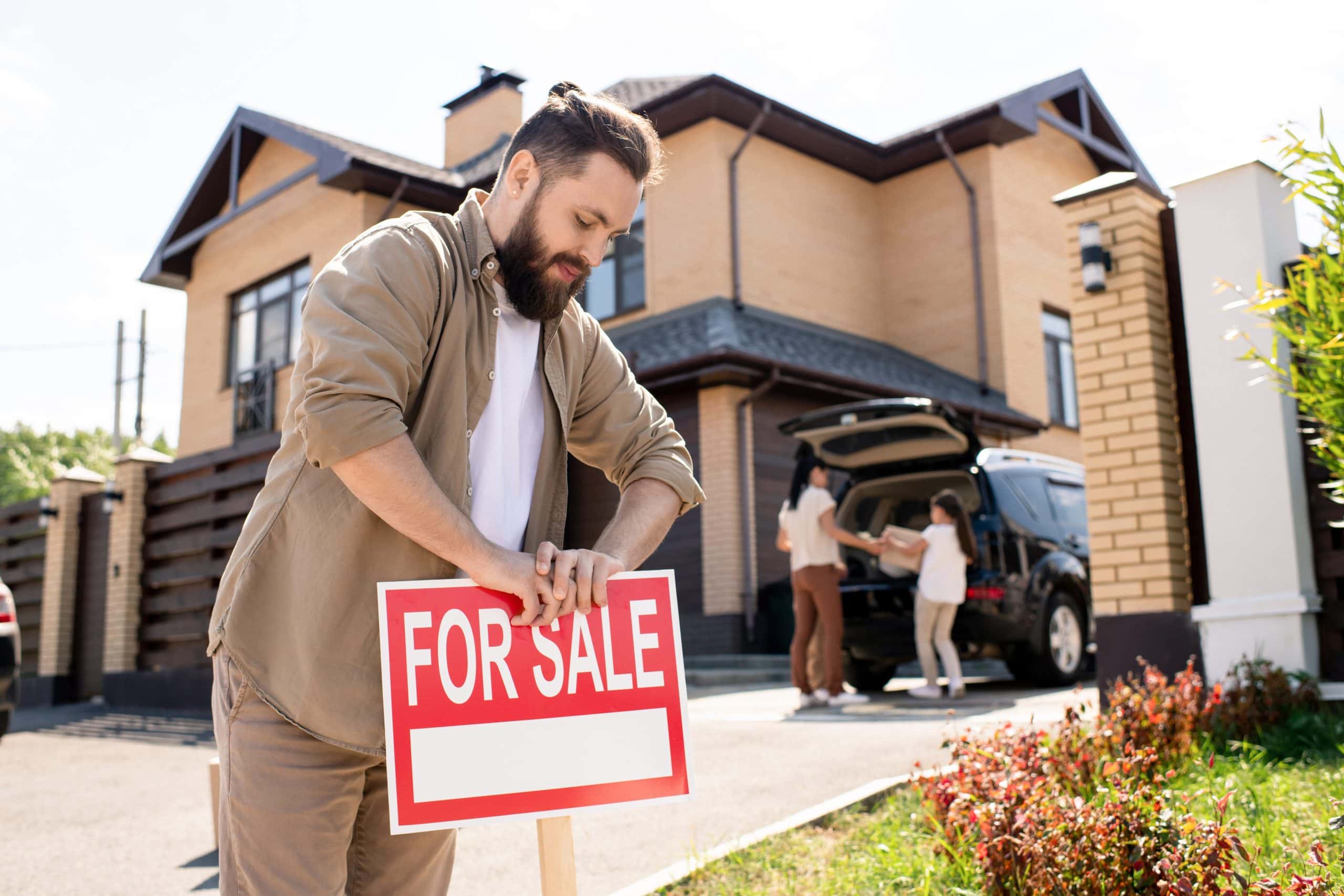 Man placing a For Sale sign in front of a modern brick house in Calgary while a family loads their car in the driveway — symbolizing moving, real estate listings, and homeownership transitions.