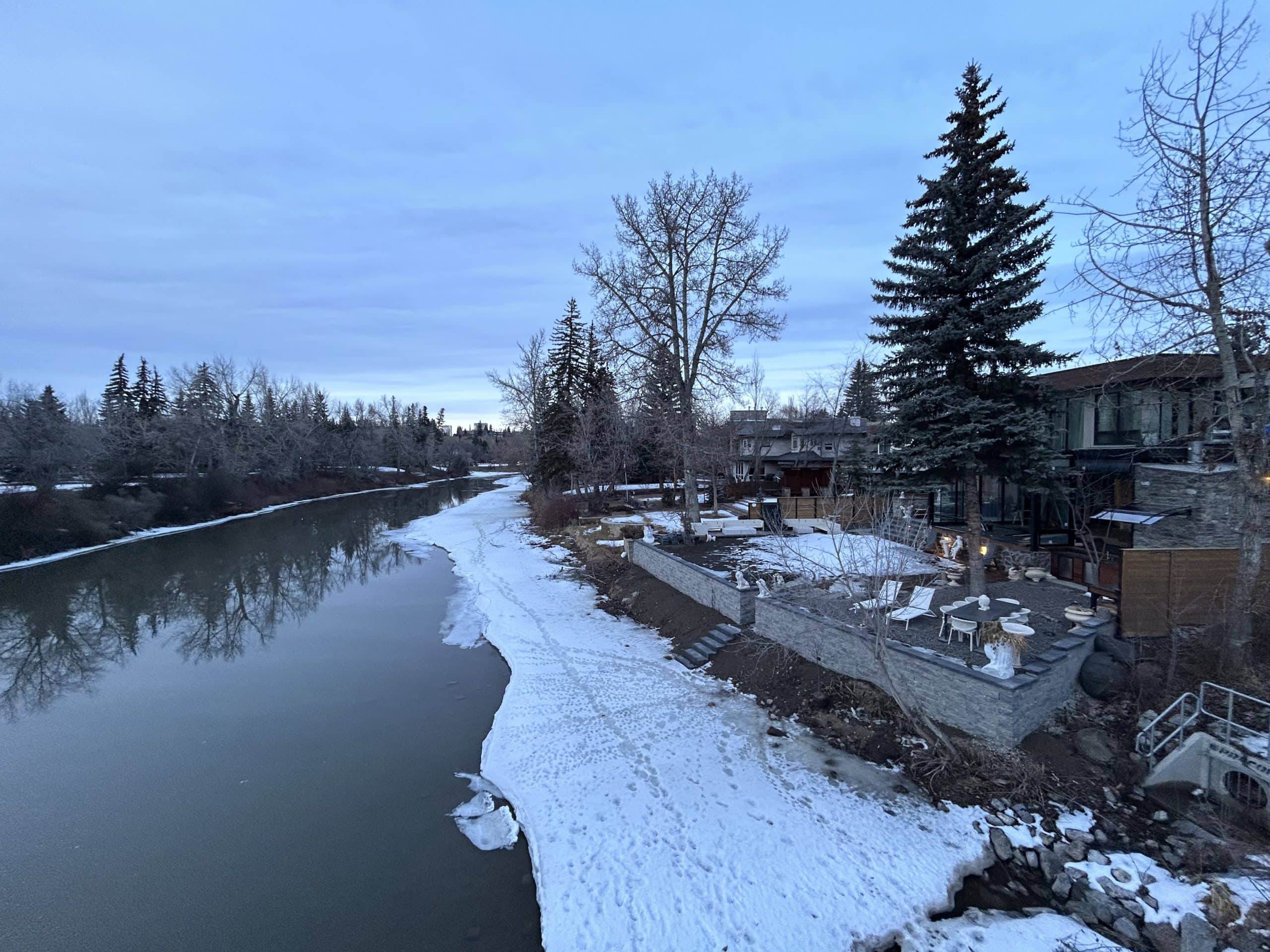 Winter view of Calgary’s Elbow River with modern riverside homes, snow-covered banks, and evergreen trees at dusk.