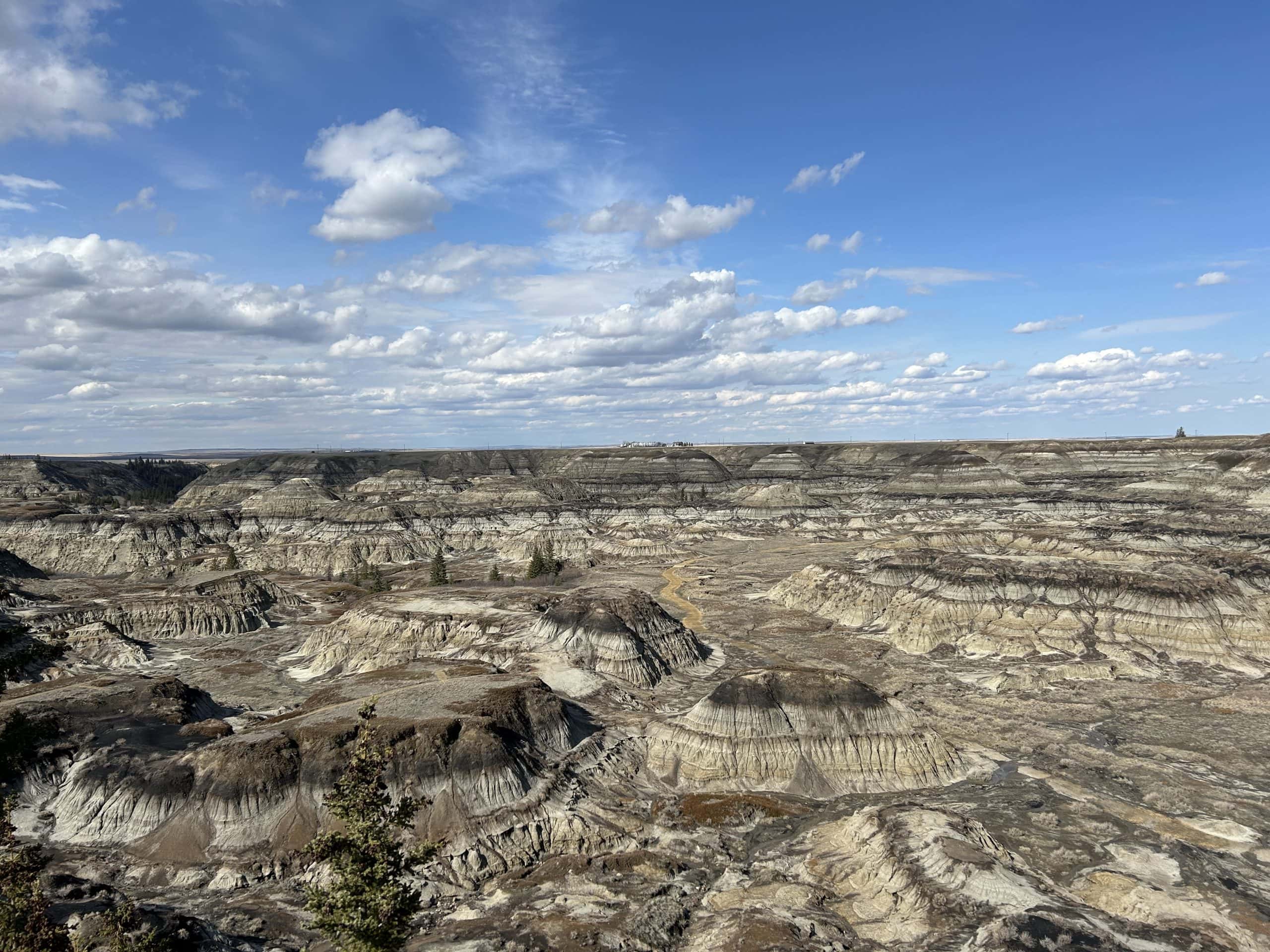 Expansive view of the Drumheller Badlands in Alberta, showcasing layered rock formations, deep canyons, and striking desert-like terrain under a partly cloudy blue sky.