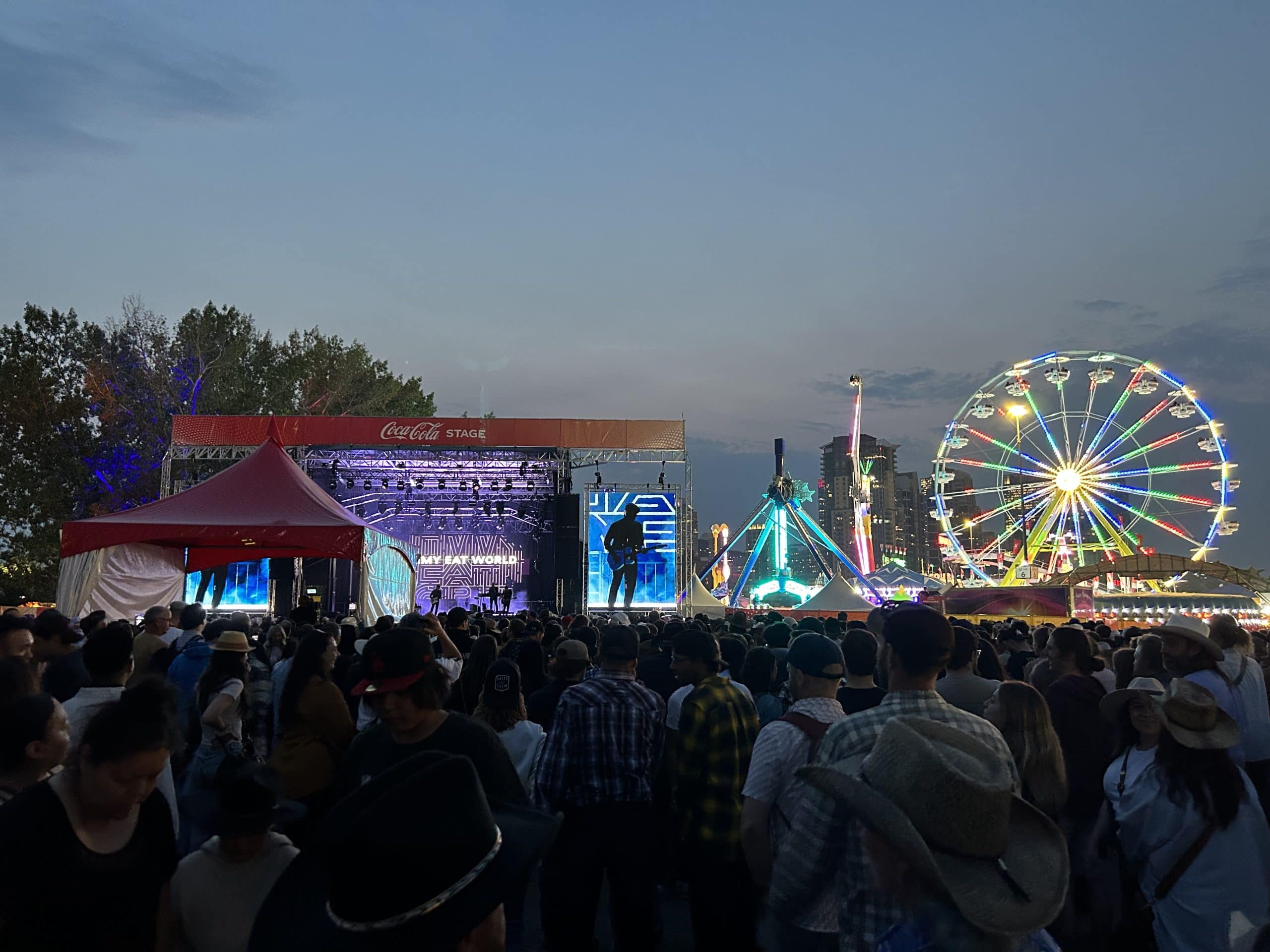 Evening crowd enjoying a live concert at the Calgary Stampede midway, featuring the Coca-Cola Stage, colourful lights, a Ferris wheel, and a festive summer atmosphere under the twilight sky.