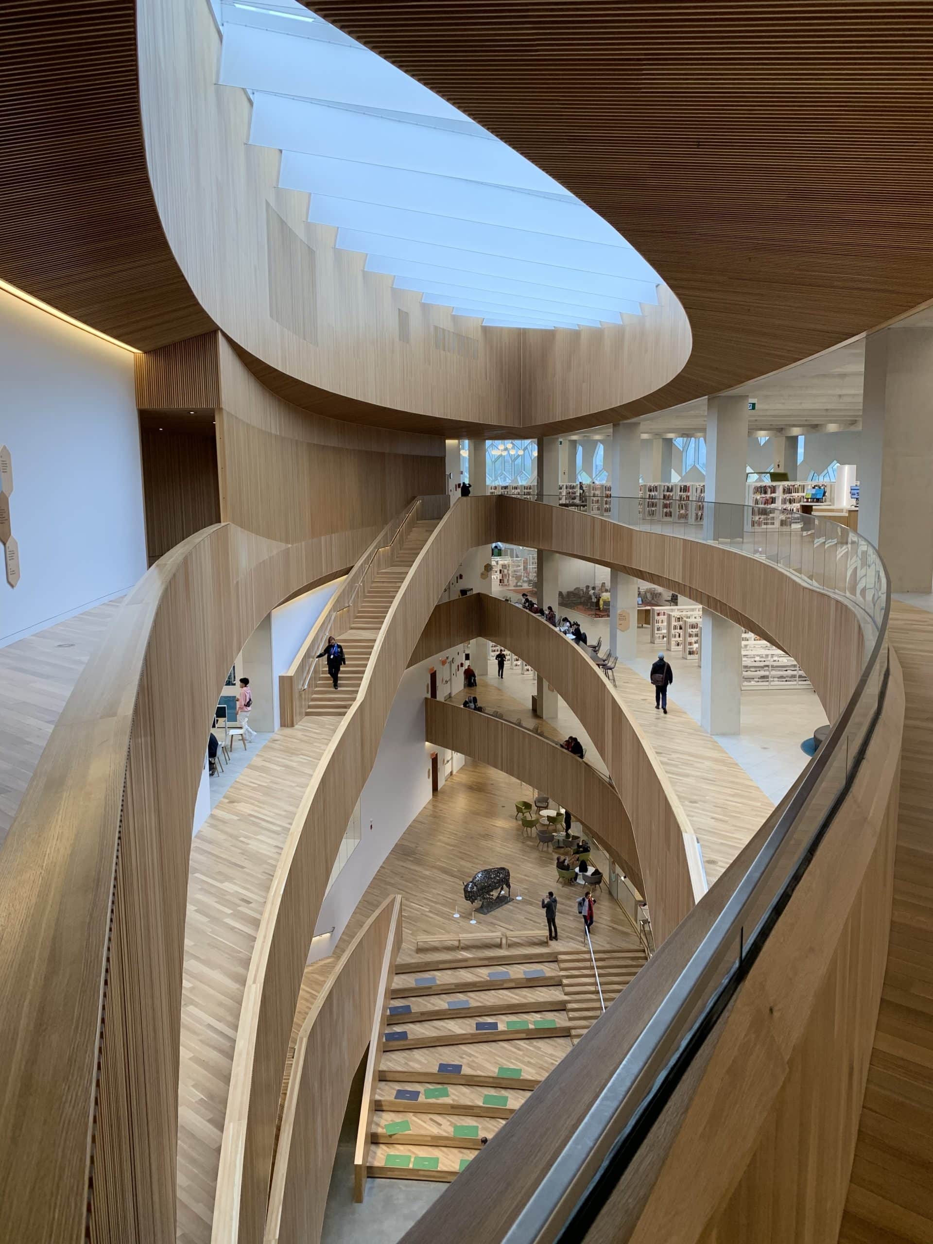 Interior view of the Calgary Central Library showcasing its stunning modern architecture, featuring curved wooden walkways, open spaces, natural light from skylights, and multi-level design with people exploring the vibrant public space.