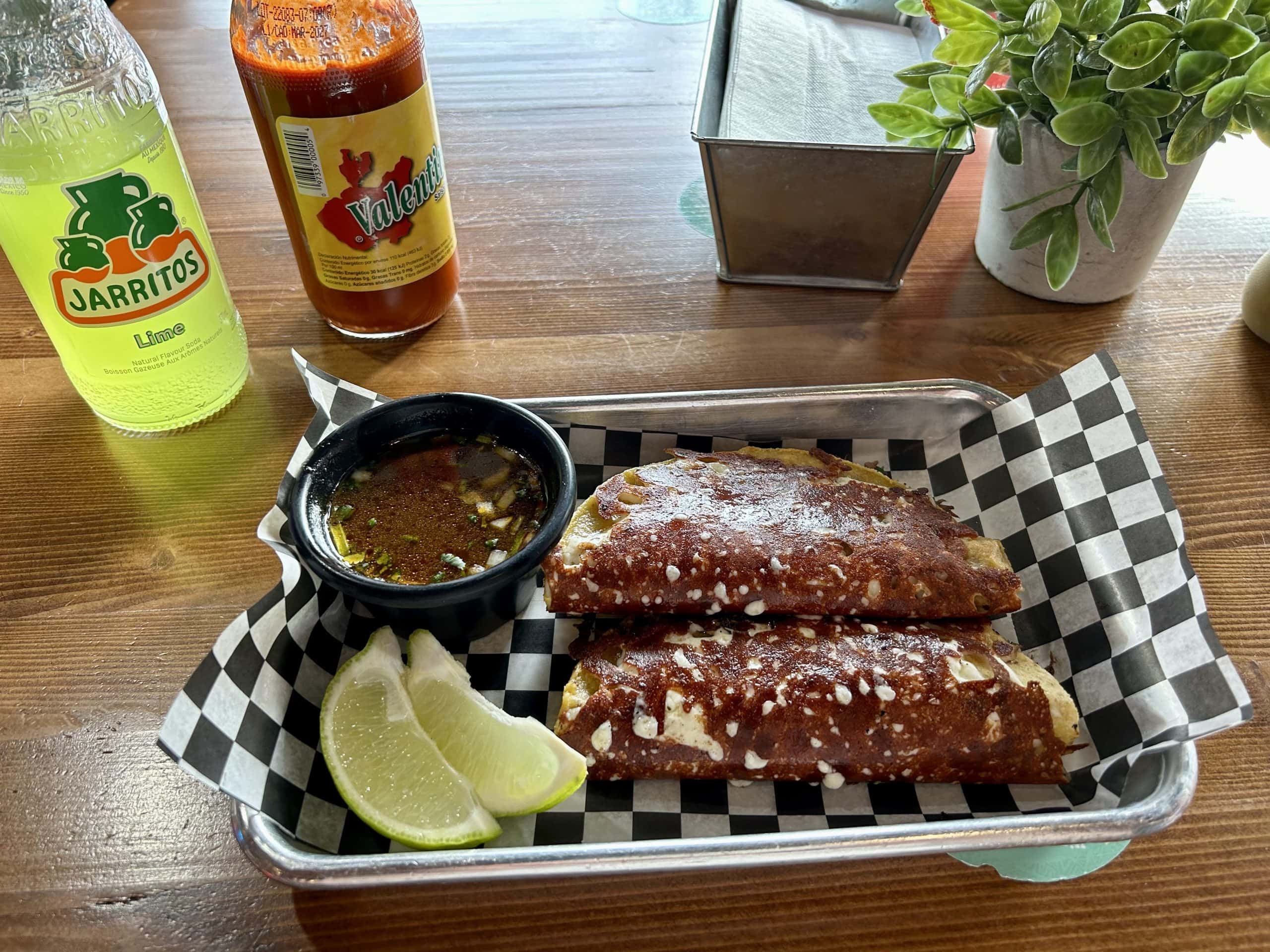 Close-up of two birria tacos served with dipping broth, lime wedges, and a bottle of Jarritos lime soda on a wooden table, creating a vibrant and authentic Mexican street food setting at Tu Taco Calgary.