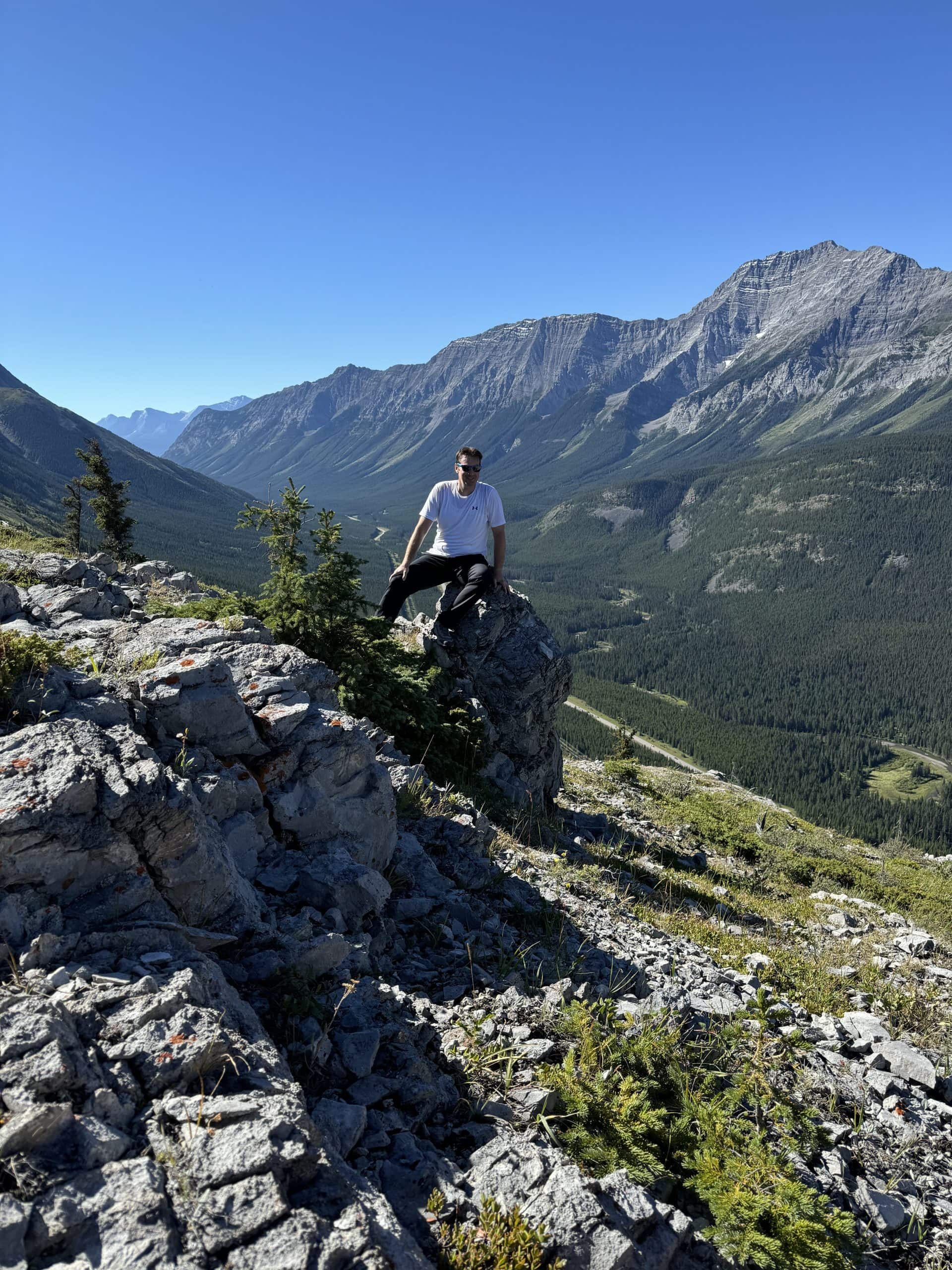 Hiker resting on a rocky ridge in Kananaskis Country, Alberta, surrounded by towering mountain peaks, deep forested valleys, and clear blue skies on a perfect summer day.