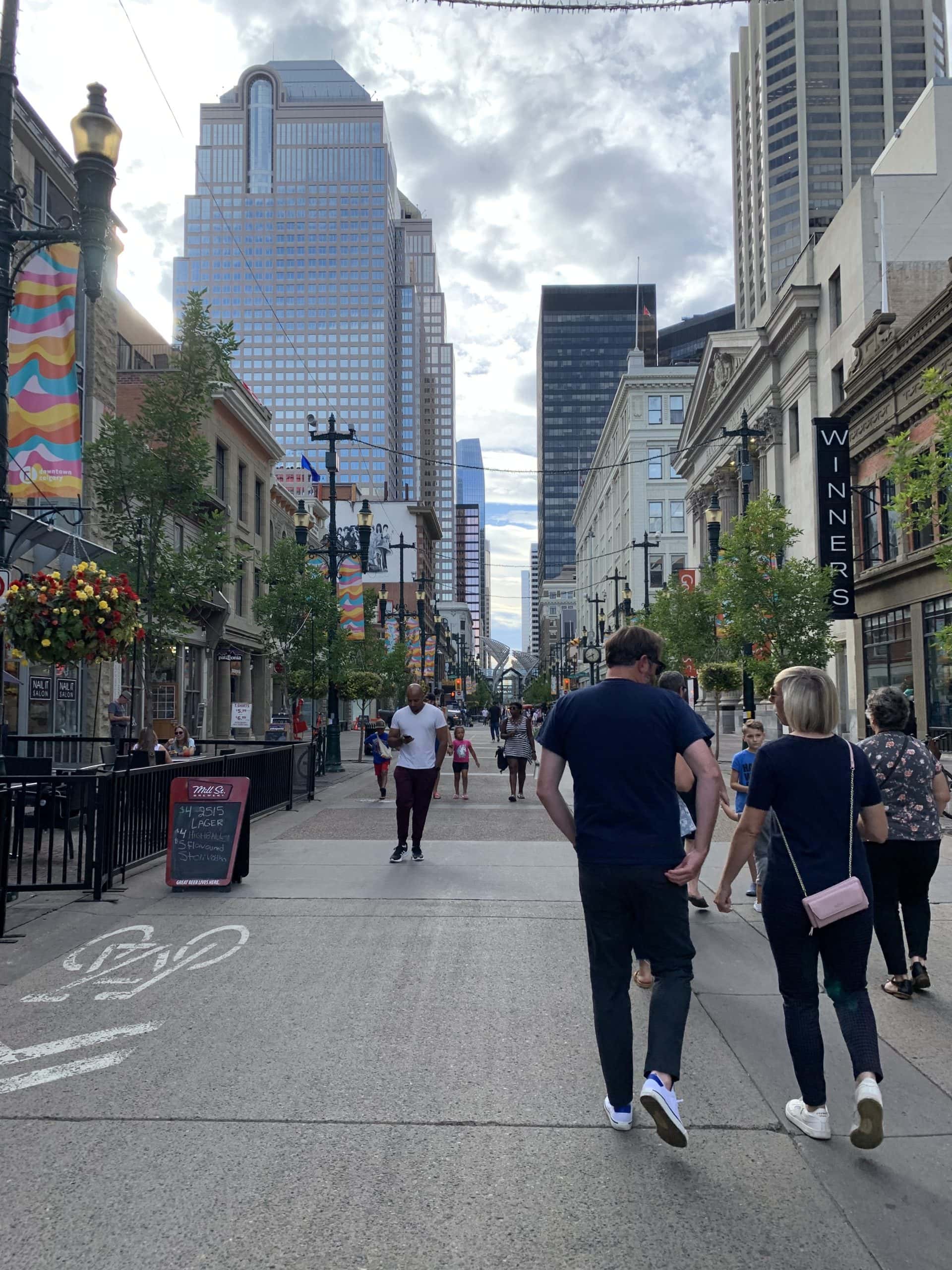 Pedestrians enjoying a summer day along Stephen Avenue in downtown Calgary, surrounded by historic buildings, modern skyscrapers, street banners, and patios that highlight the city’s lively urban atmosphere.