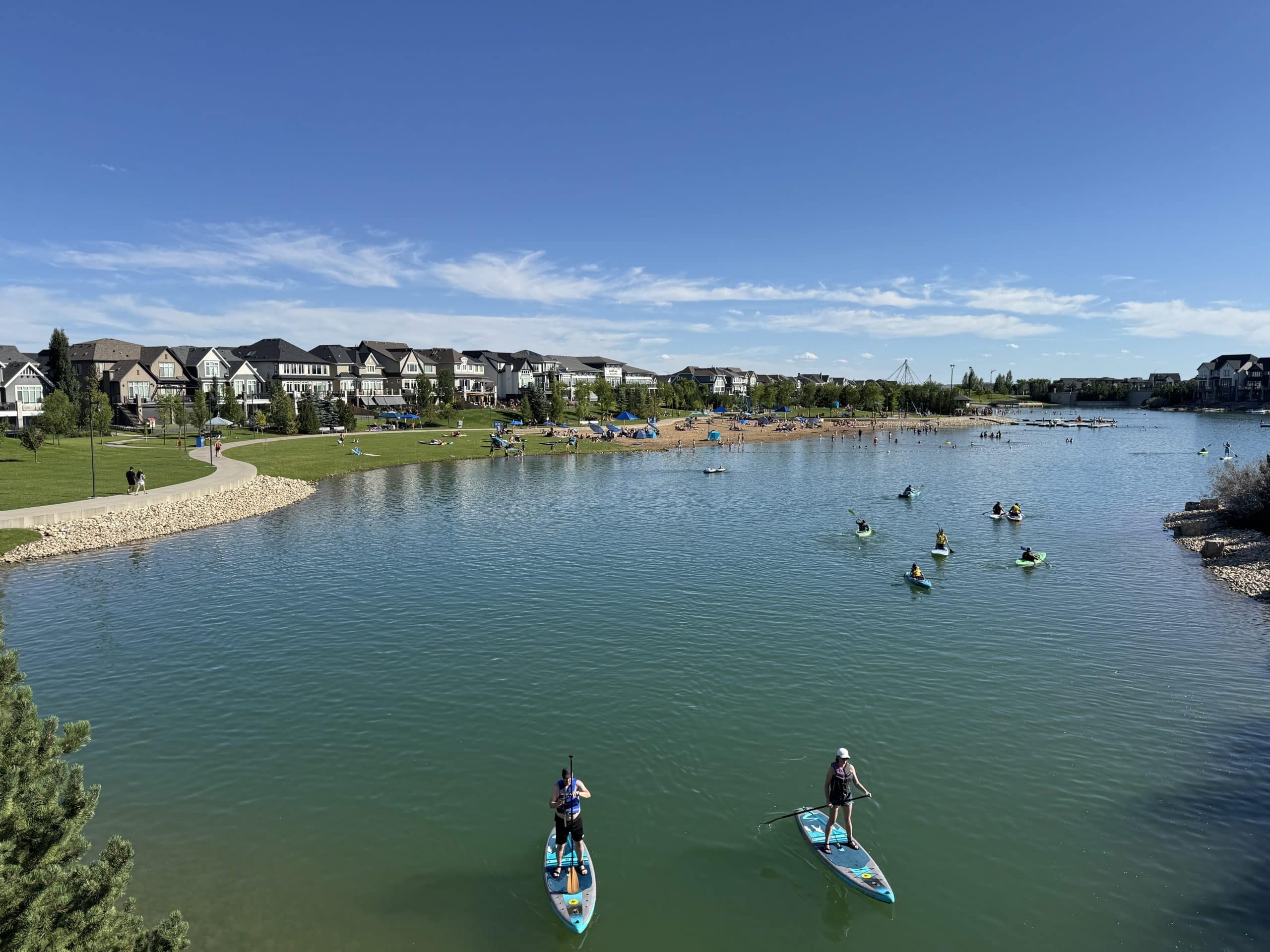 Summer day at Mahogany Lake in Calgary with people paddleboarding, kayaking, and relaxing on the sandy beach, surrounded by modern lakeside homes and green park space under a bright blue sky.