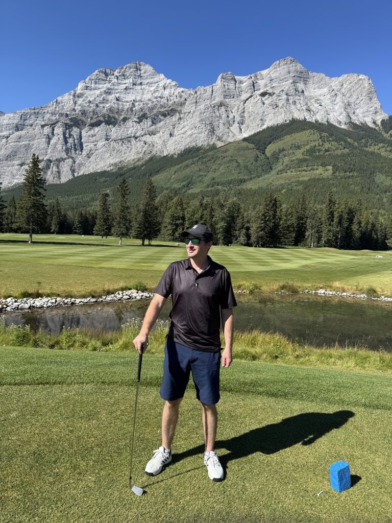 Golfer standing on a scenic fairway in Kananaskis, Alberta, surrounded by lush greens and dramatic Rocky Mountain peaks under a clear blue sky.