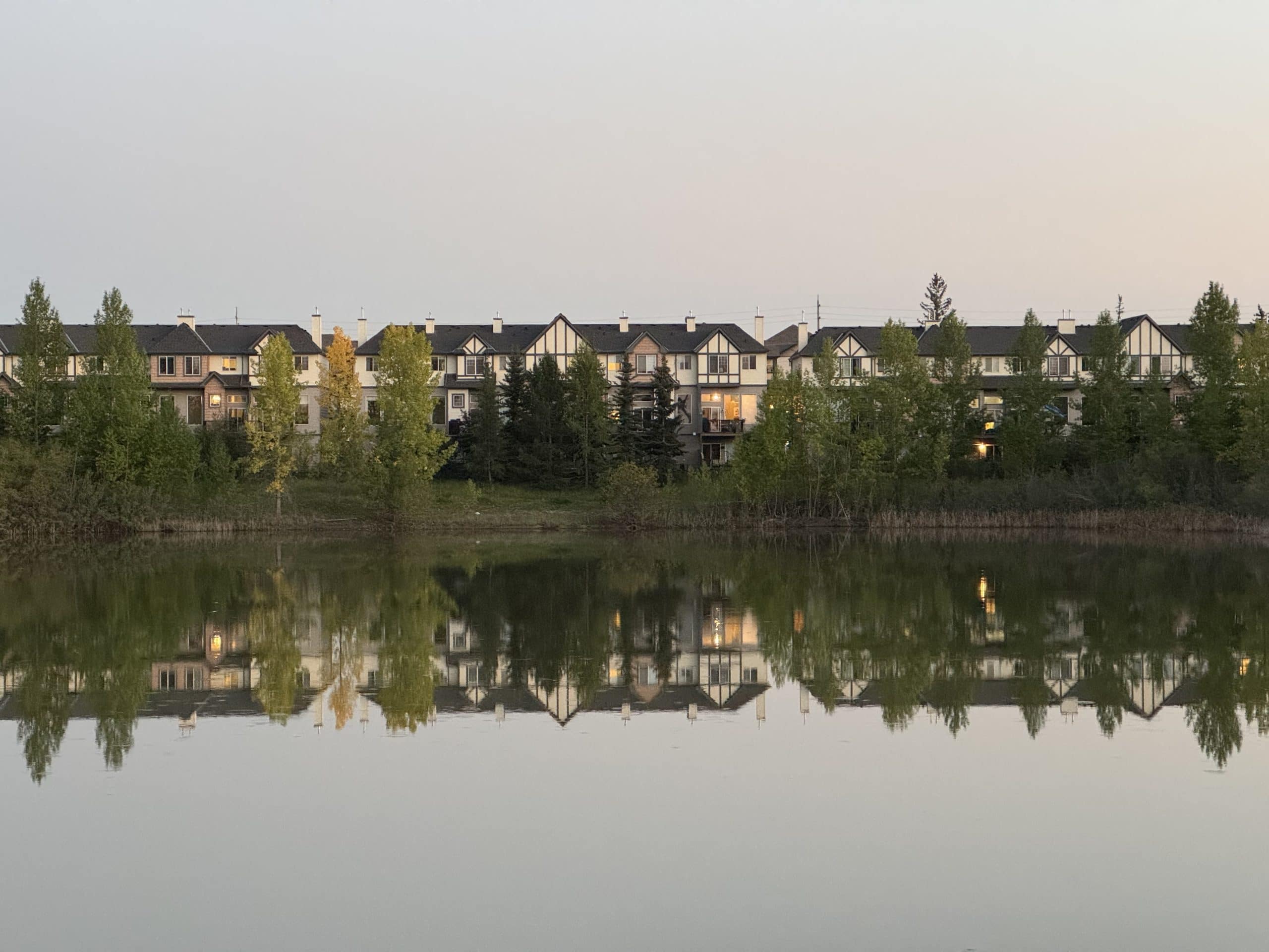 Townhomes in Evergreen Calgary reflecting on a calm pond at sunset, showcasing the peaceful scenery and natural beauty of this southwest neighbourhood.