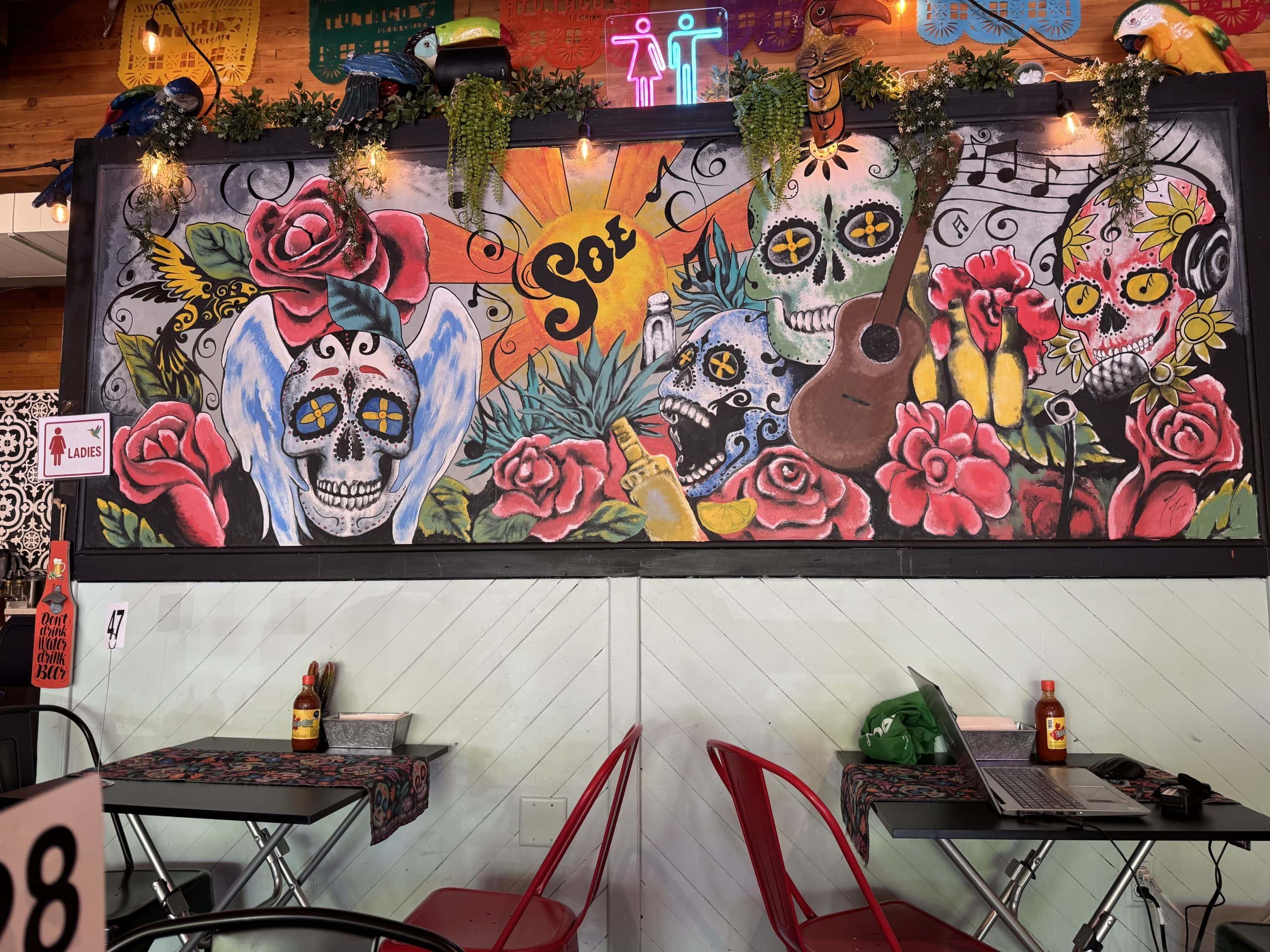 Colourful Day of the Dead mural inside Tu Taco in Calgary, featuring vibrant sugar skulls, roses, and musical motifs, surrounded by hanging plants, festive lighting, and a cozy, artistic dining atmosphere.
