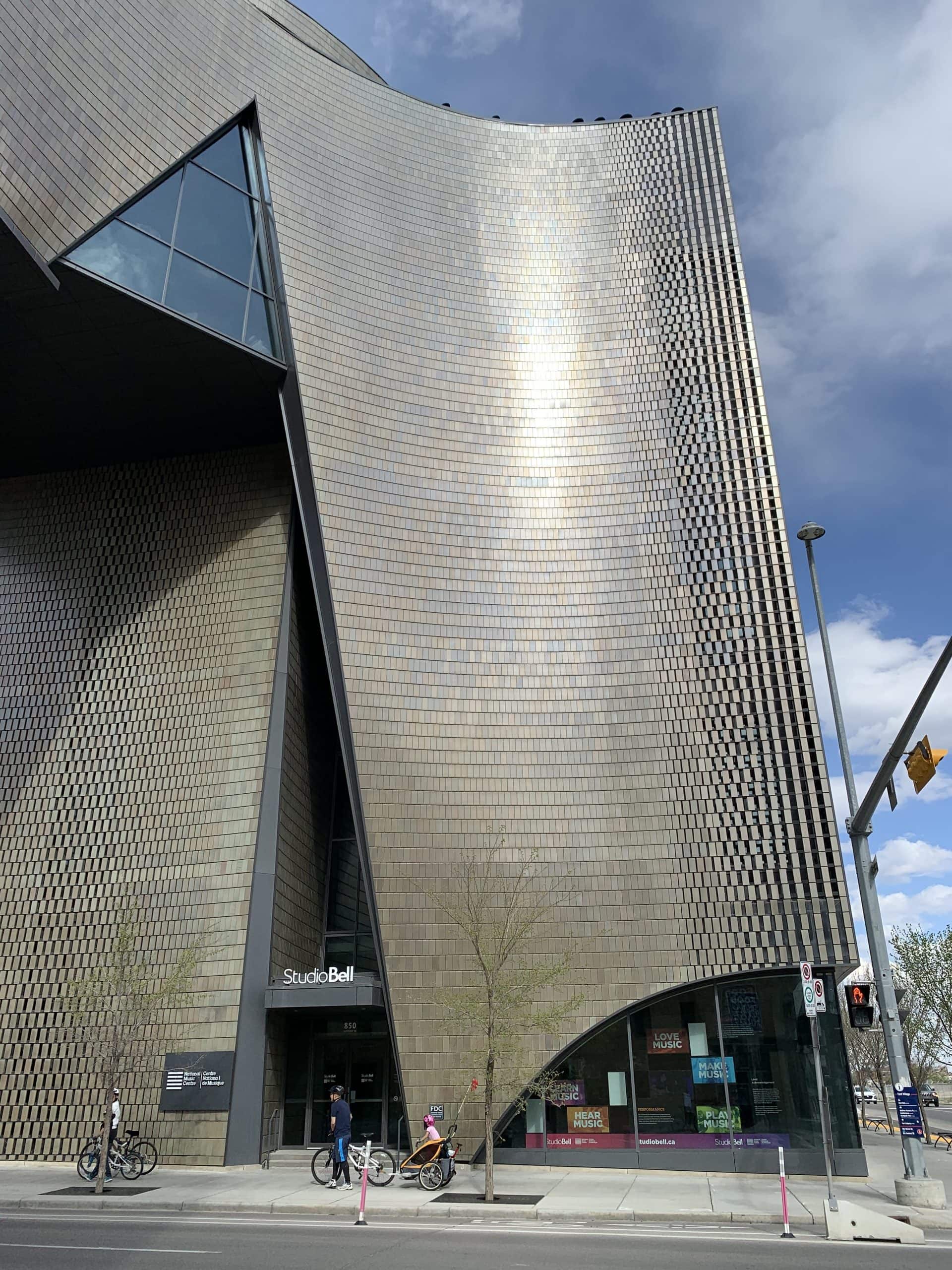 Exterior view of Studio Bell, home of the National Music Centre in Calgary, showcasing its striking modern architecture with shimmering curved tiles, geometric windows, and pedestrians passing by on a sunny day.