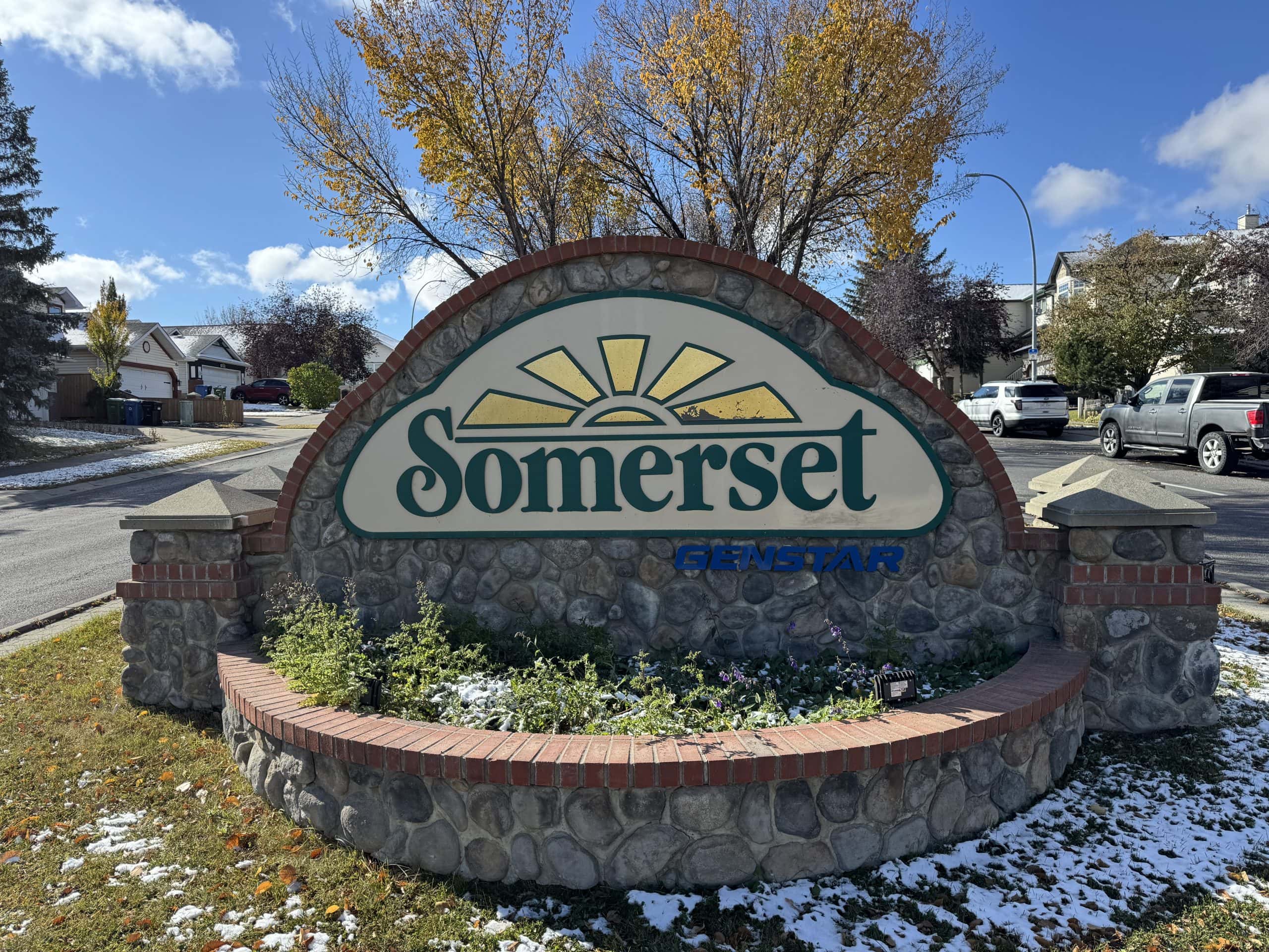 Stone entrance sign for the Somerset neighbourhood in southwest Calgary featuring a sun logo, autumn trees, residential streets, and a light dusting of early season snow.