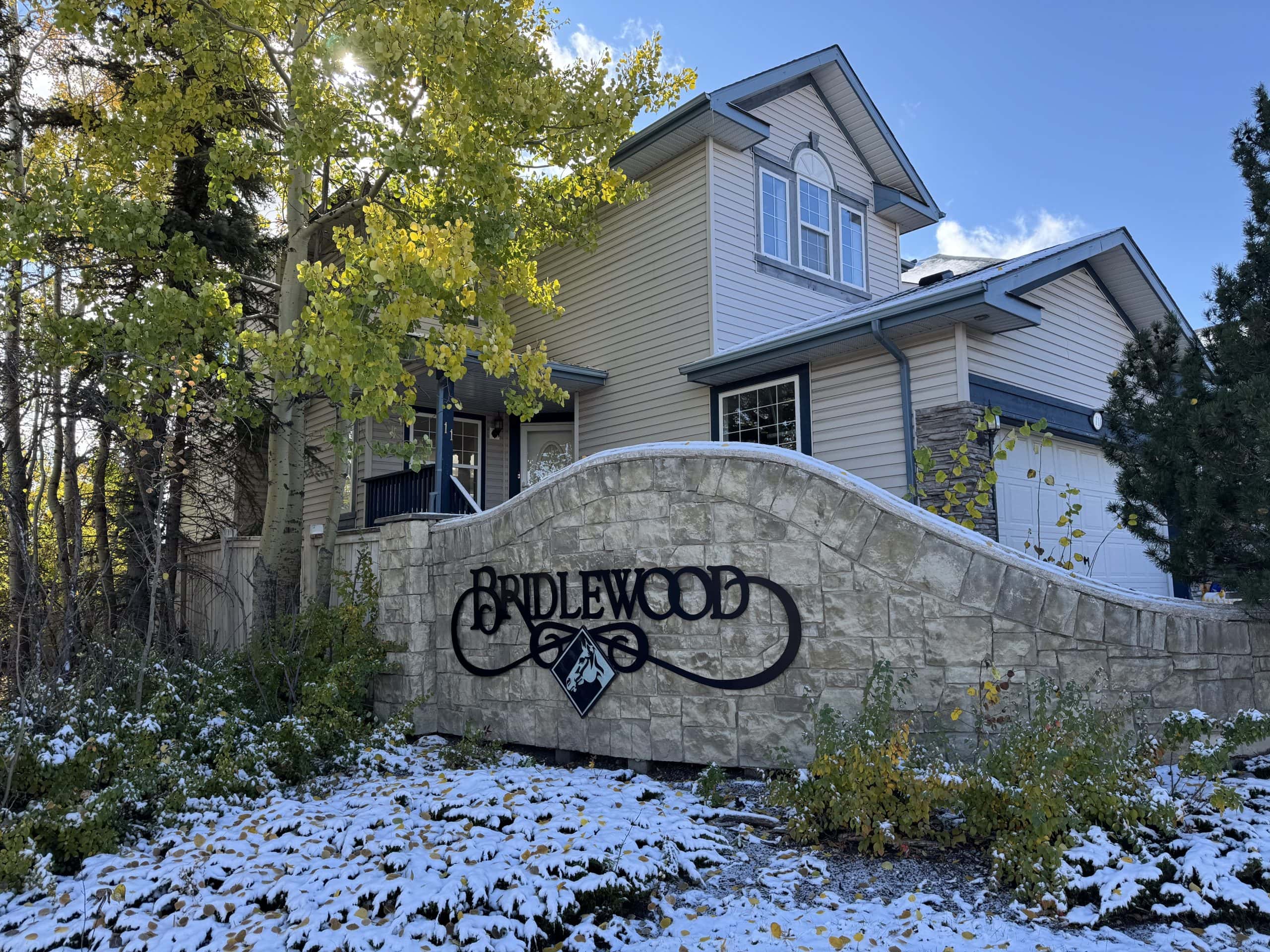 Bridlewood Calgary neighbourhood entrance sign set against mature trees, light snow on the ground, and a two storey suburban home in the background on a sunny autumn day.