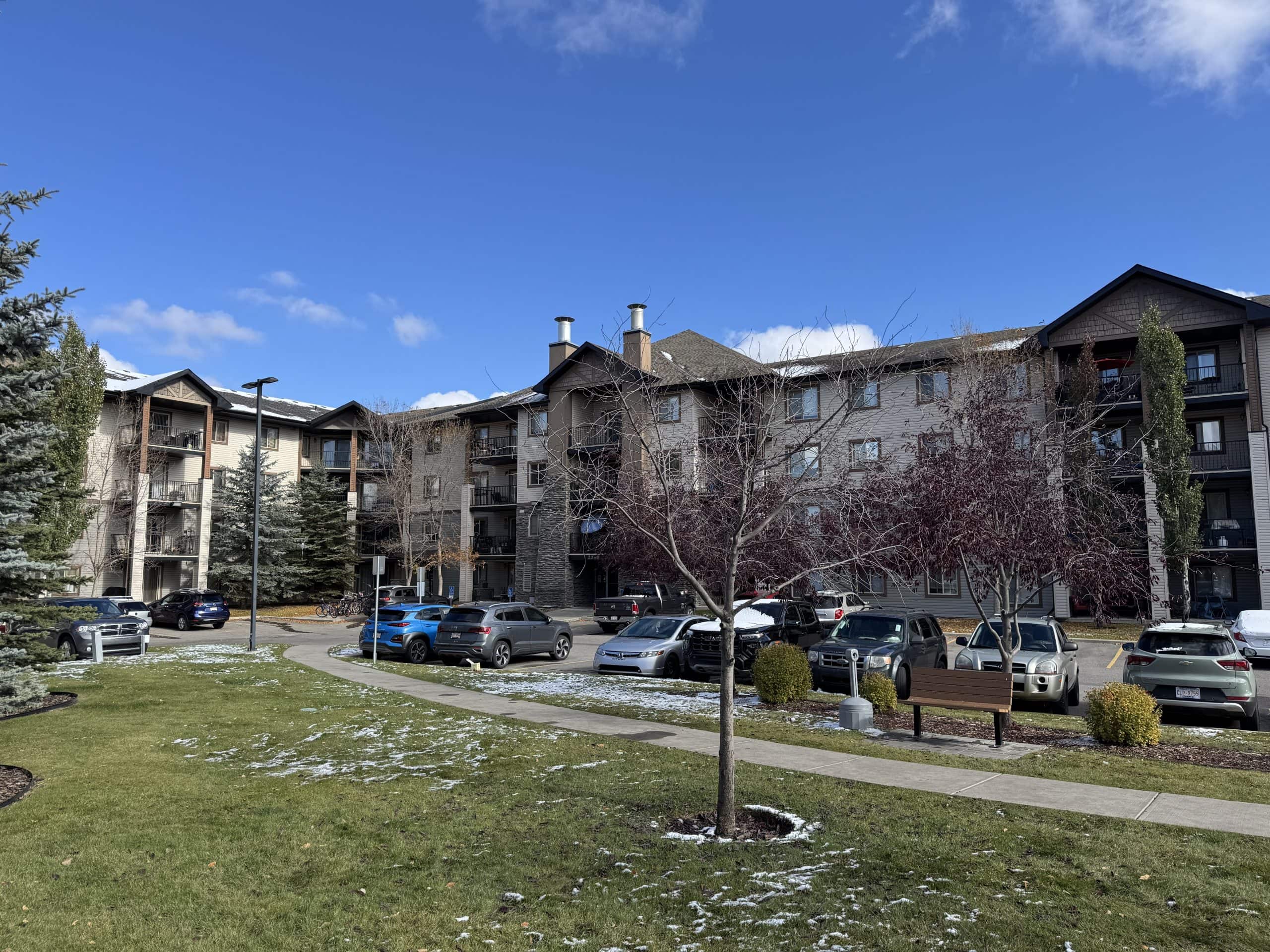 Four storey Bridlewood condo buildings in southwest Calgary with balconies, mature trees, surface parking, and light early season snow on the ground under a bright blue sky.