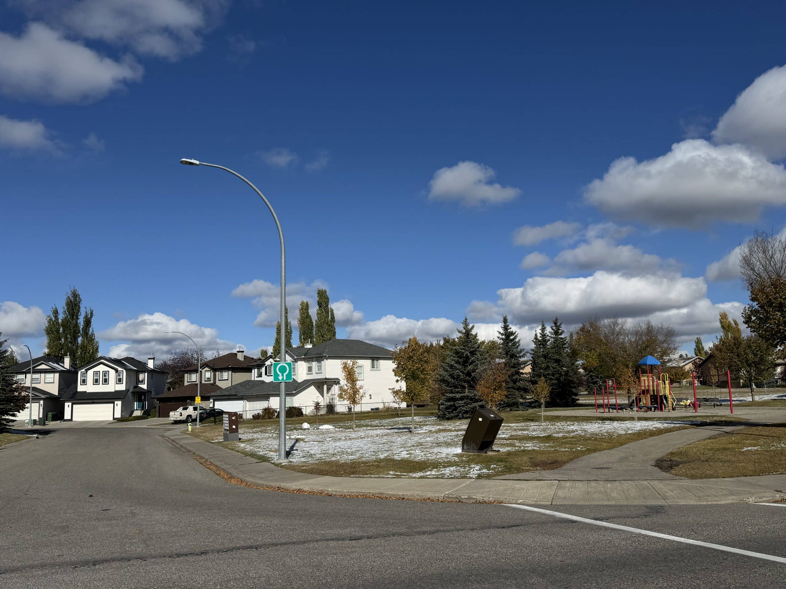 Neighbourhood park in Shawnessy Calgary with a playground, mature trees, and nearby single family homes under a bright blue sky on a crisp fall day.