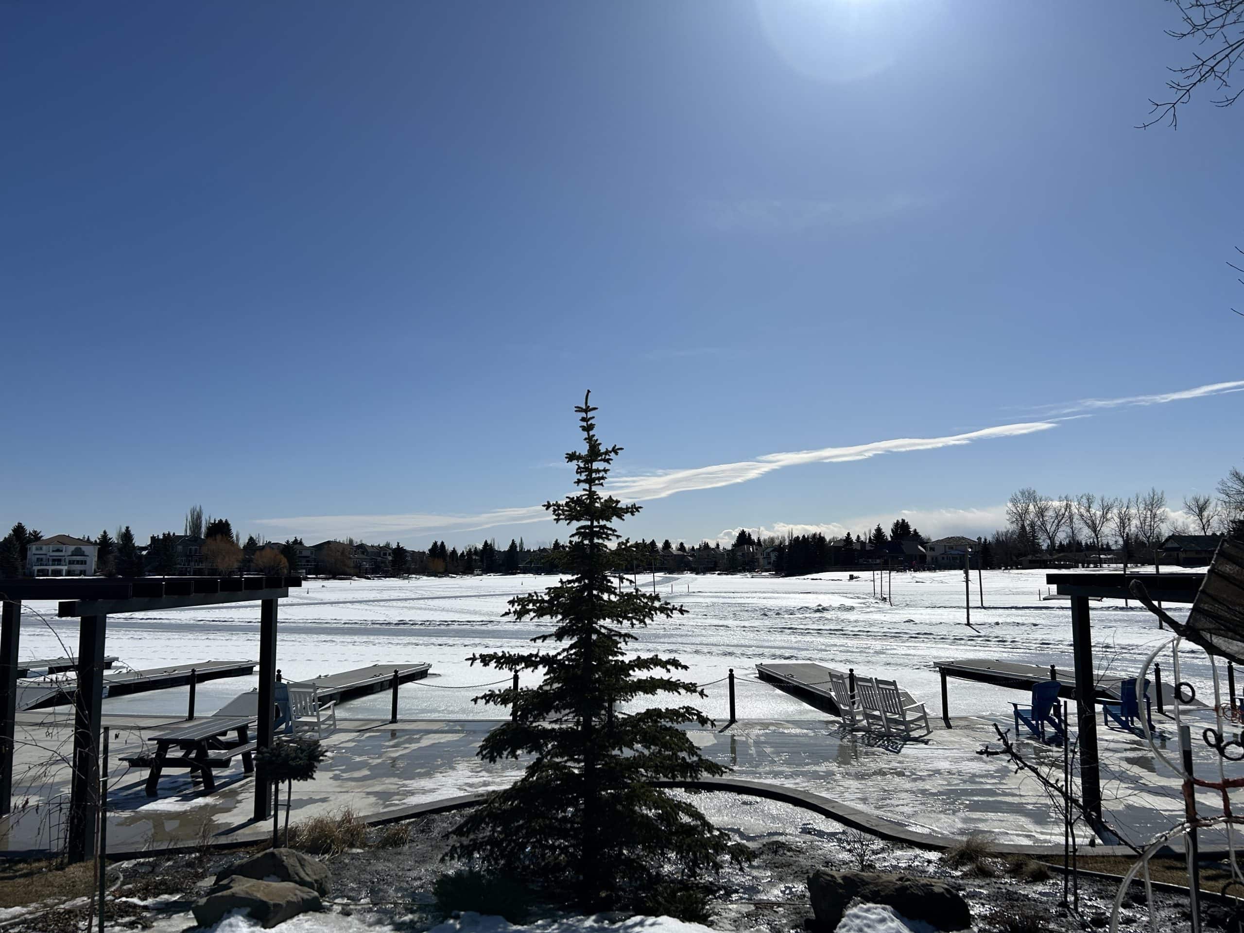 Frozen Lake Sundance in Calgary on a bright winter day with docks, Adirondack chairs, and surrounding lakeside homes under a clear blue sky.