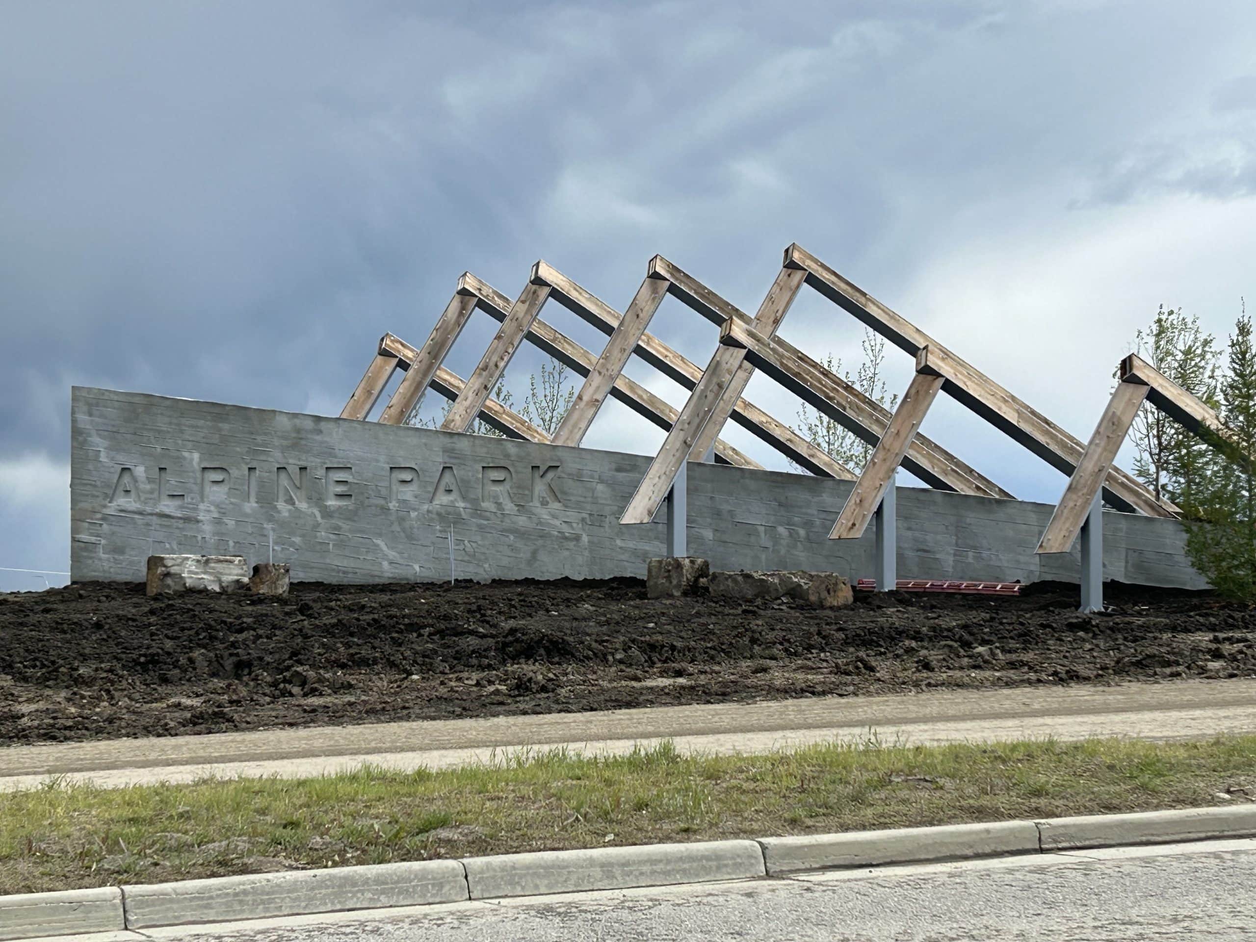 Entrance sign for Alpine Park in southwest Calgary with modern wooden beams and a concrete wall under a cloudy sky.
