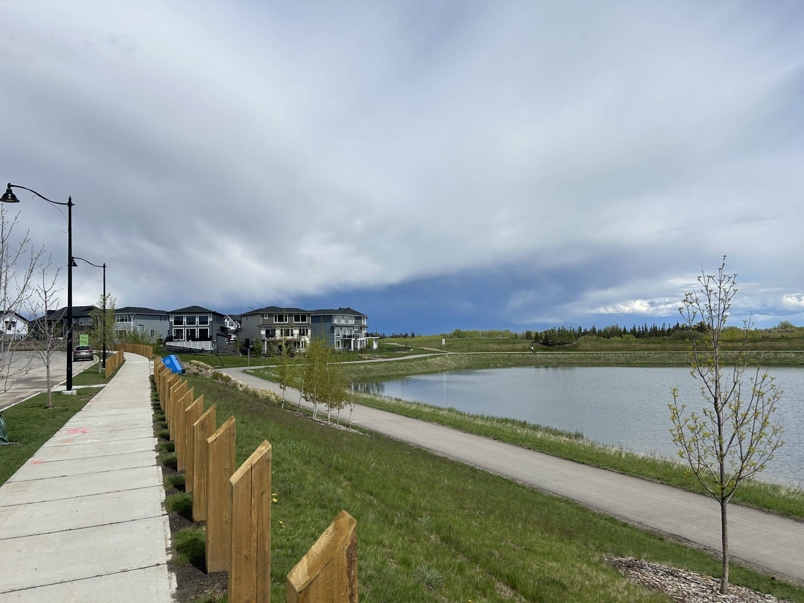 Walking path beside the storm pond in Pine Creek Calgary, with modern homes, young trees, and dramatic spring clouds over the new southwest community.