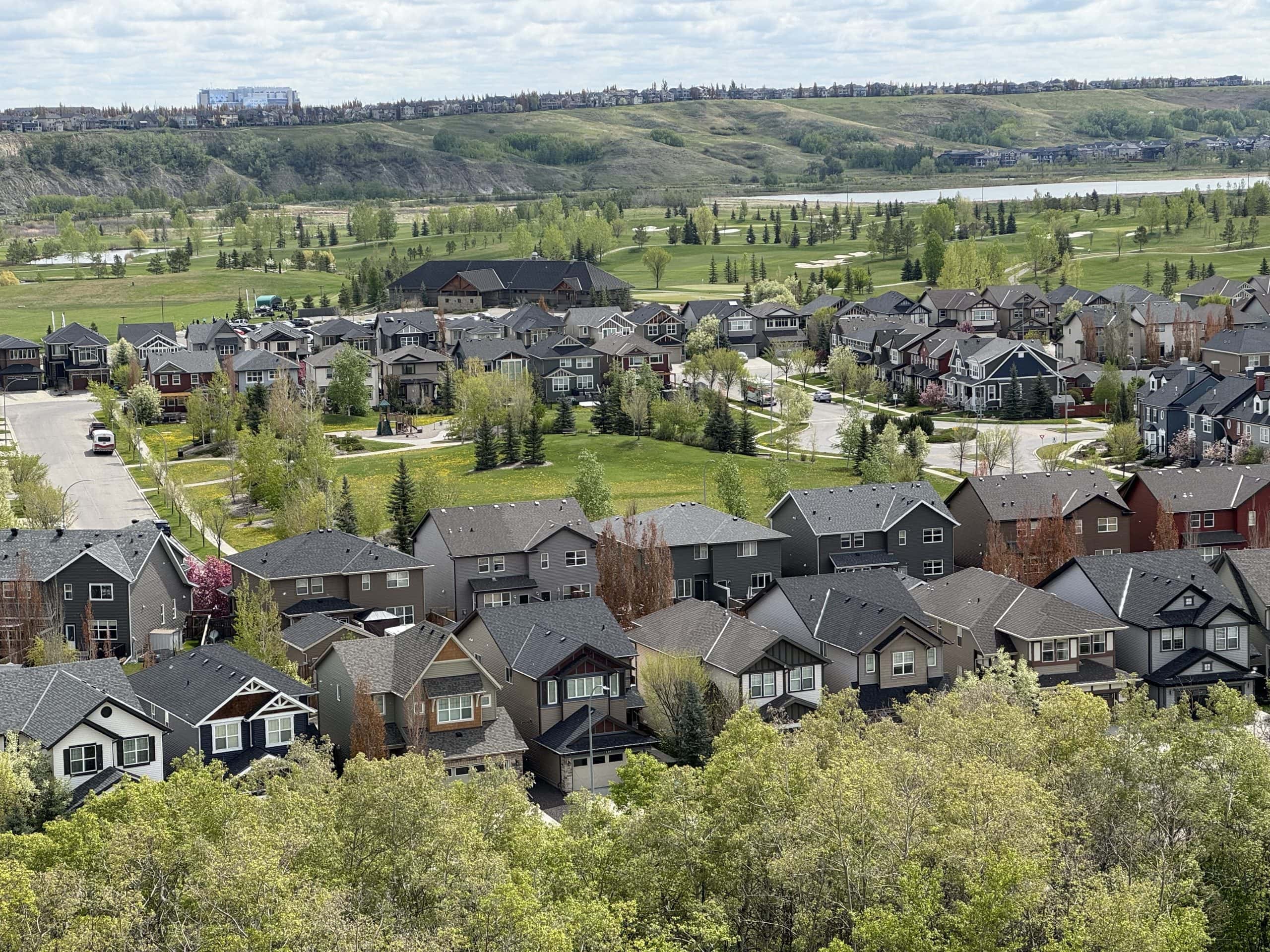 Panoramic view of a Calgary suburban neighbourhood with modern homes, tree lined streets, open green space, and a golf course and river valley in the background.