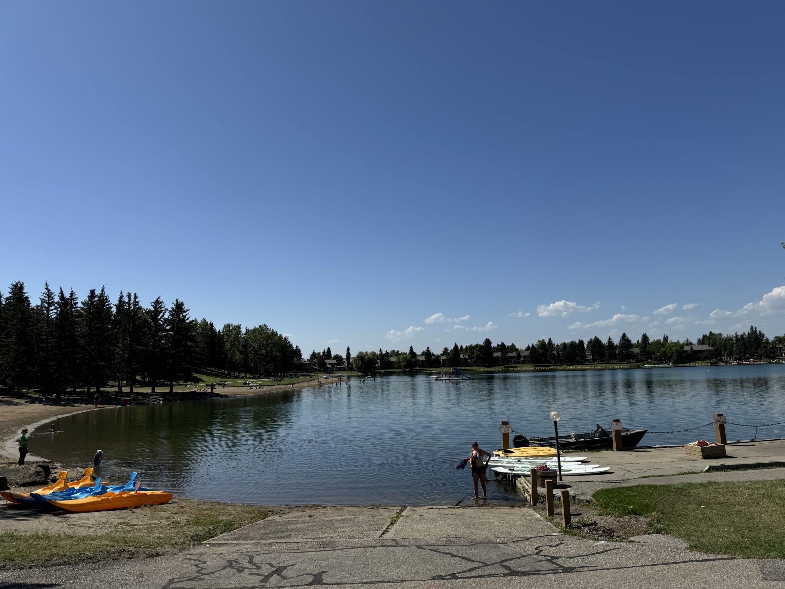 Midnapore Lake in Calgary with a sandy beach, calm water, paddleboards, kayaks, and residents enjoying the lake on a sunny summer day surrounded by mature trees and lakeside homes.