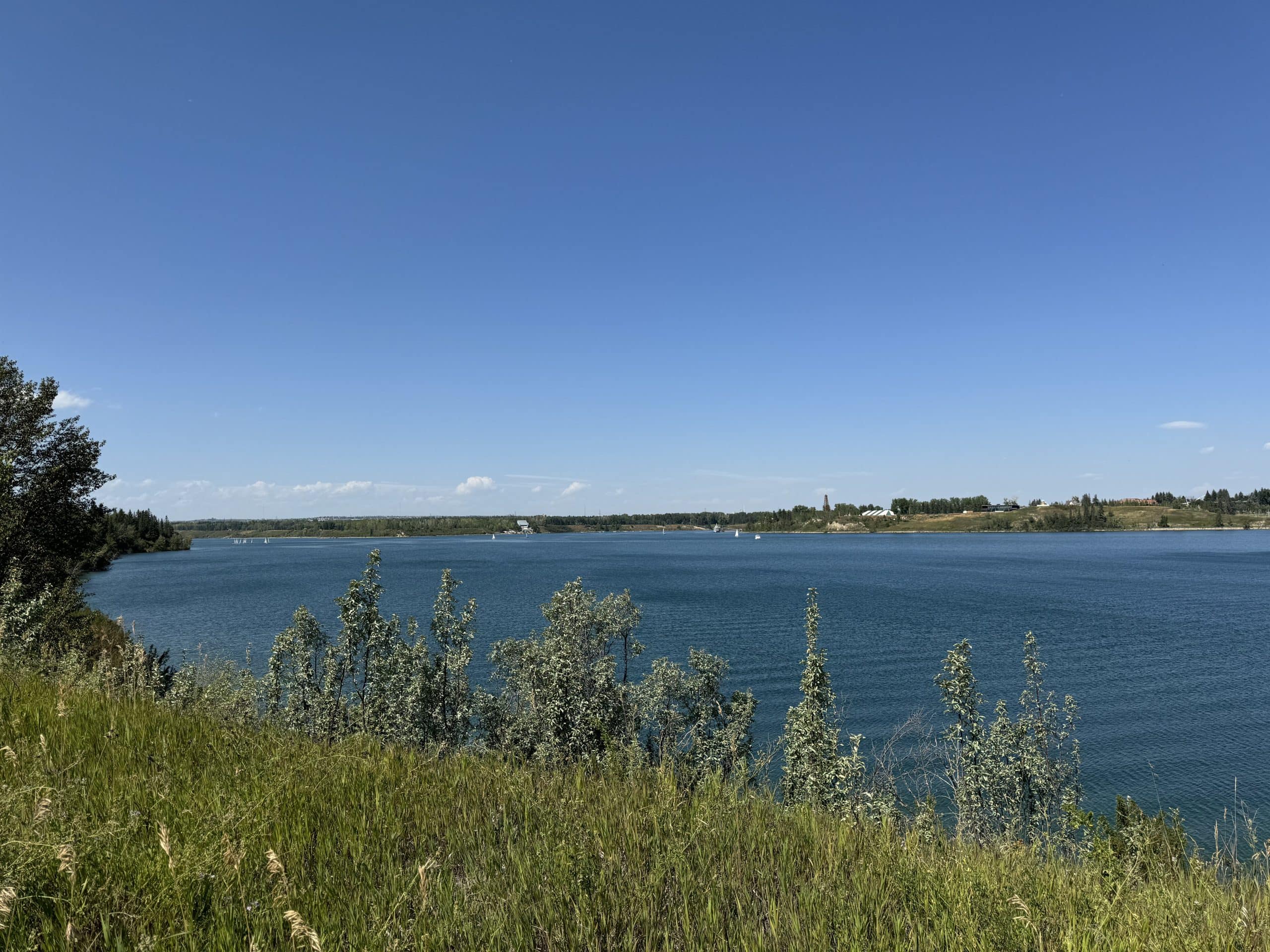 View of the Glenmore Reservoir from Bayview with clear blue water, sailboats in the distance, and green shoreline vegetation on a bright summer day in South Calgary.