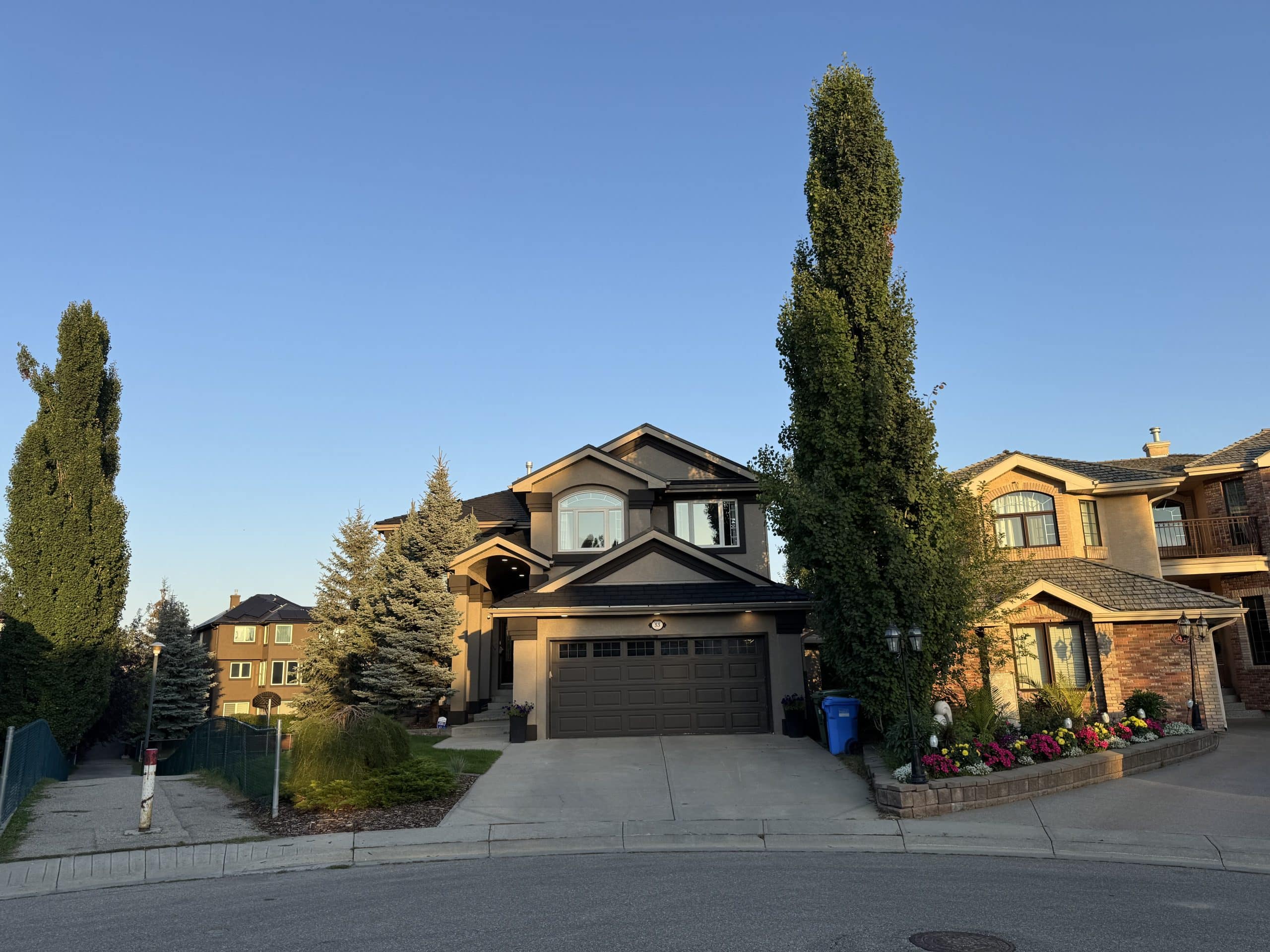 Two storey home in Evergreen Calgary with a wide driveway, tall trees, and evening sunlight highlighting the quiet suburban street.