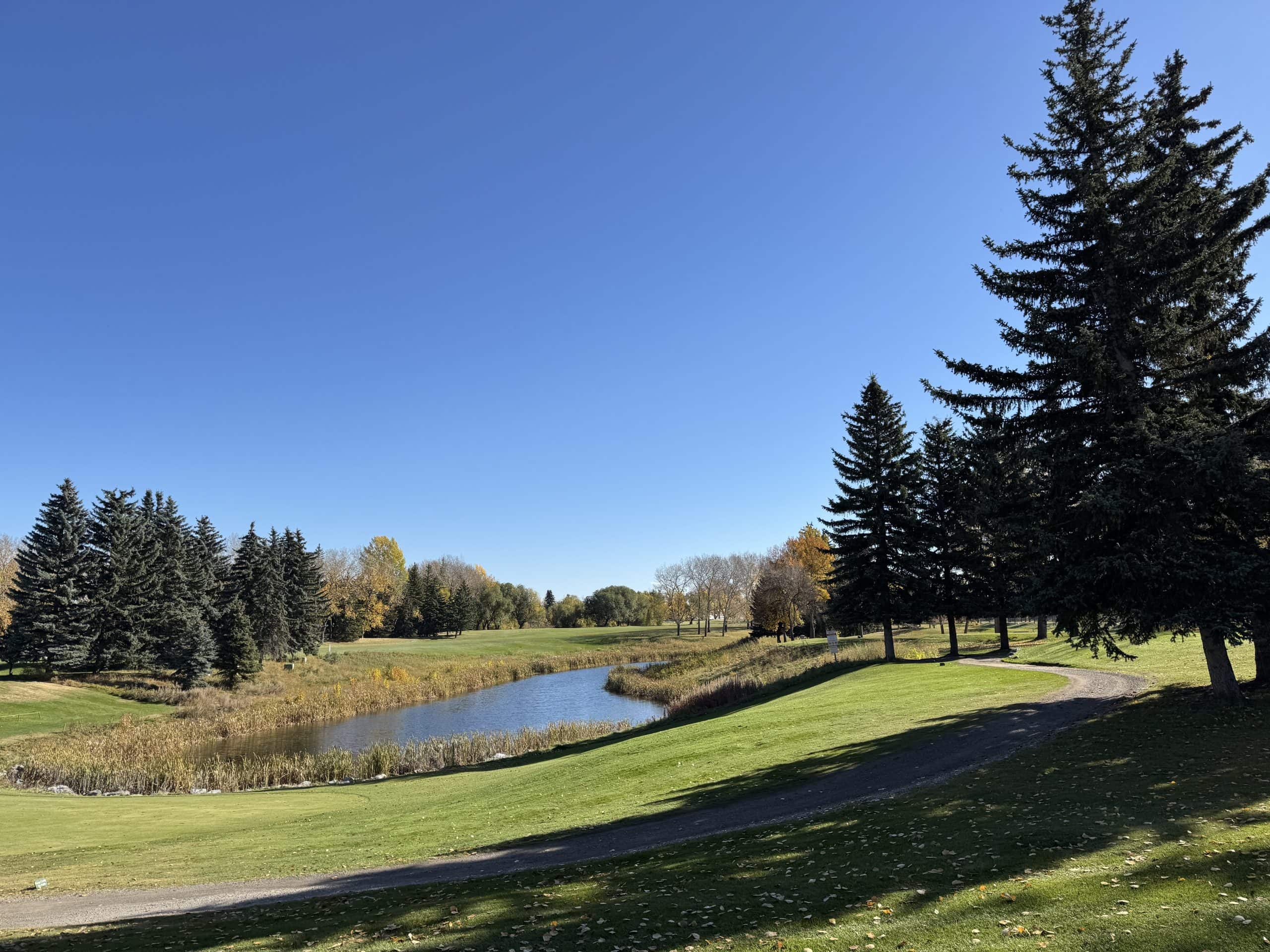 Maple Ridge Golf Course with green fairways, a small water feature, mature evergreens, and bright fall colours under a clear blue sky.