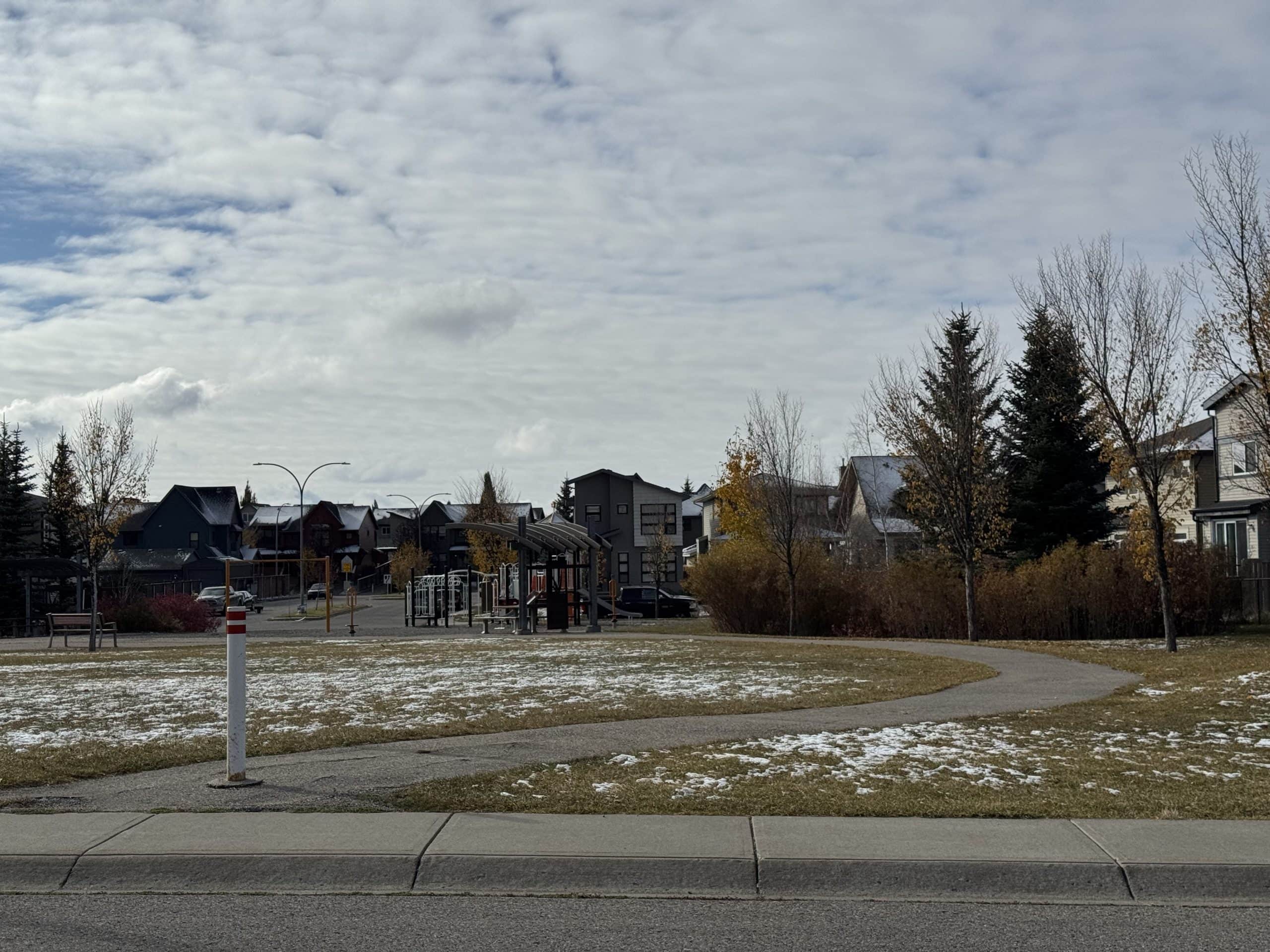Neighbourhood park in Walden Calgary with a playground, winding pathway, and modern homes in the background on a light snowy autumn day.
