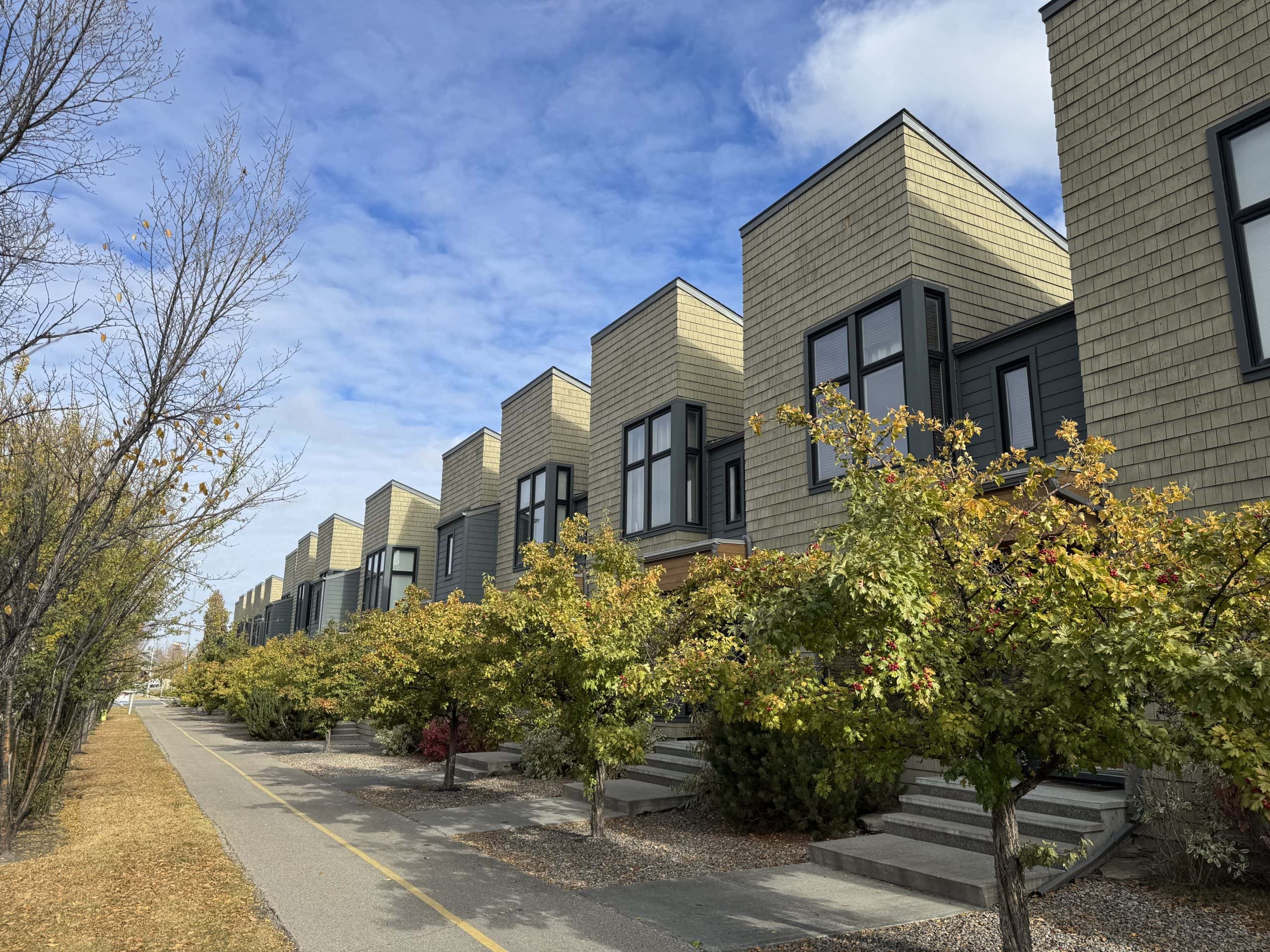 Row of modern townhomes in Walden Calgary with a tree lined walking path and autumn colours under a bright blue sky.