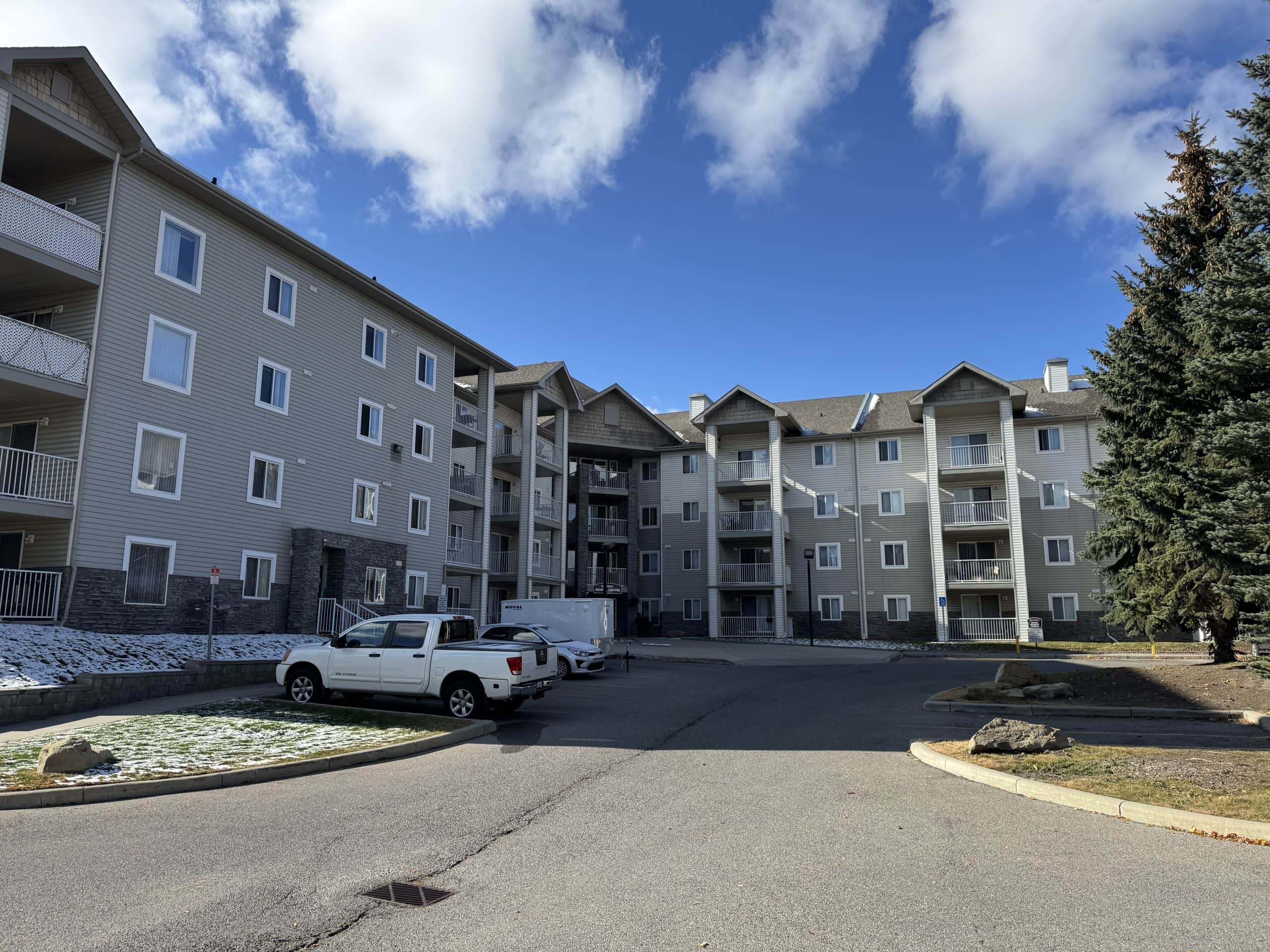 Four storey condo buildings in Somerset Calgary with covered balconies, light siding, stone accents, and a central courtyard with parking under a bright blue sky with light snow on the ground.
