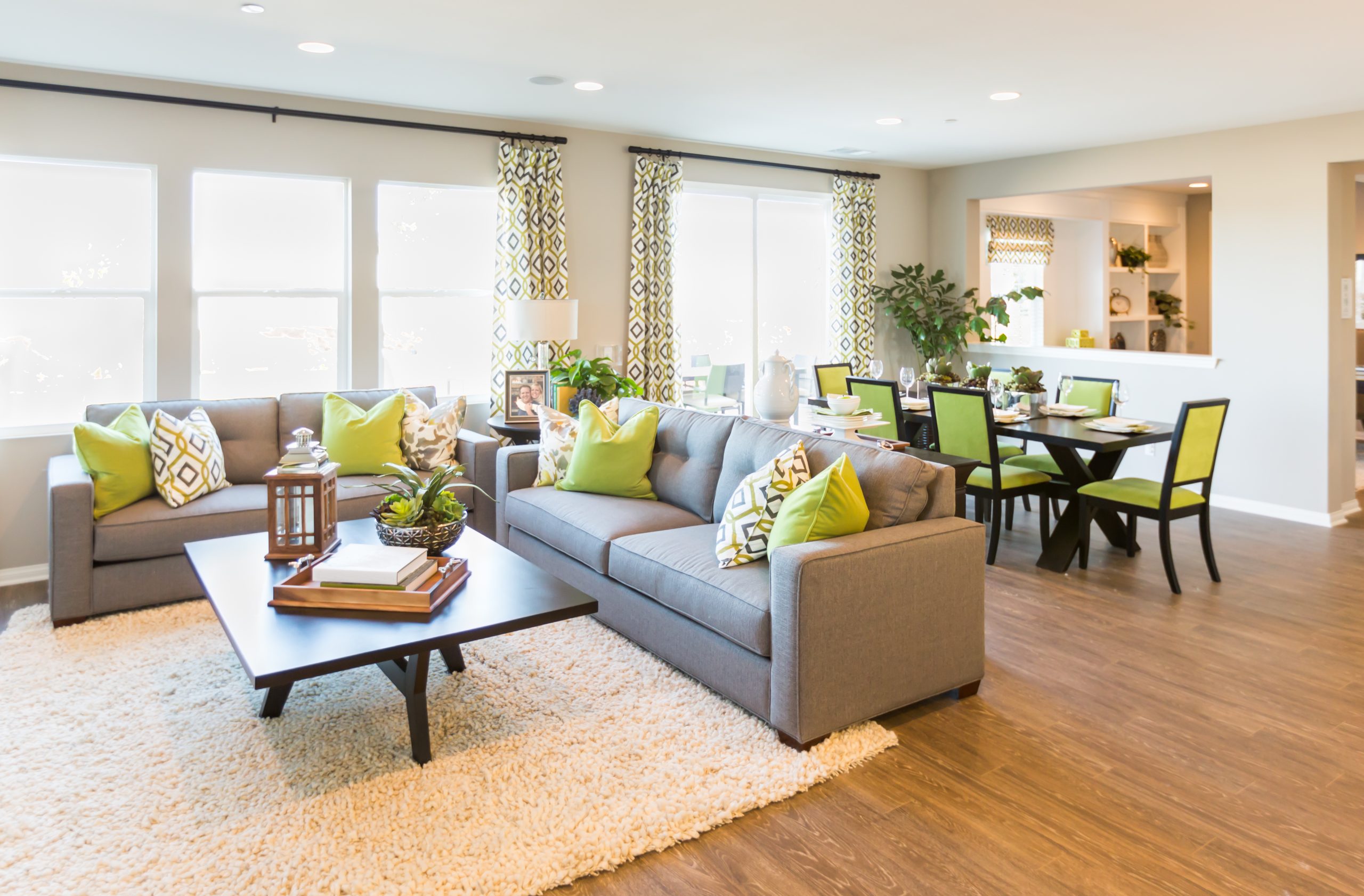 Bright open concept living and dining room with modern grey sofas, green accent pillows, hardwood floors, and large windows showcasing a clean and stylish home interior.