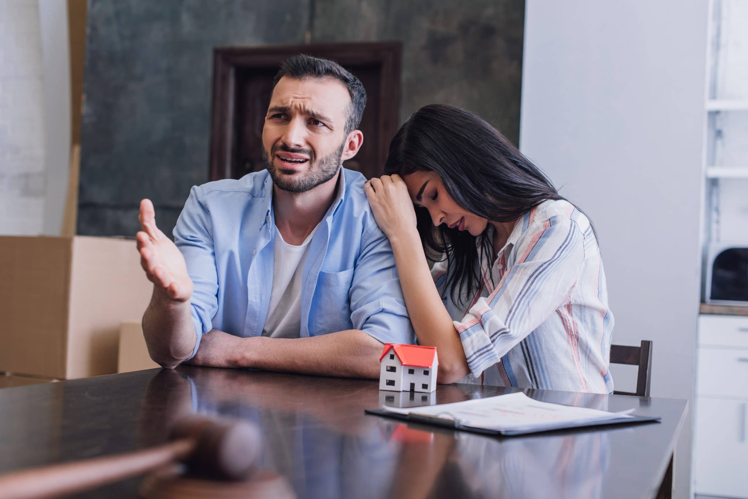 Stressed couple sitting at a table with a small house model and paperwork, showing frustration during a difficult home sale or negotiation.