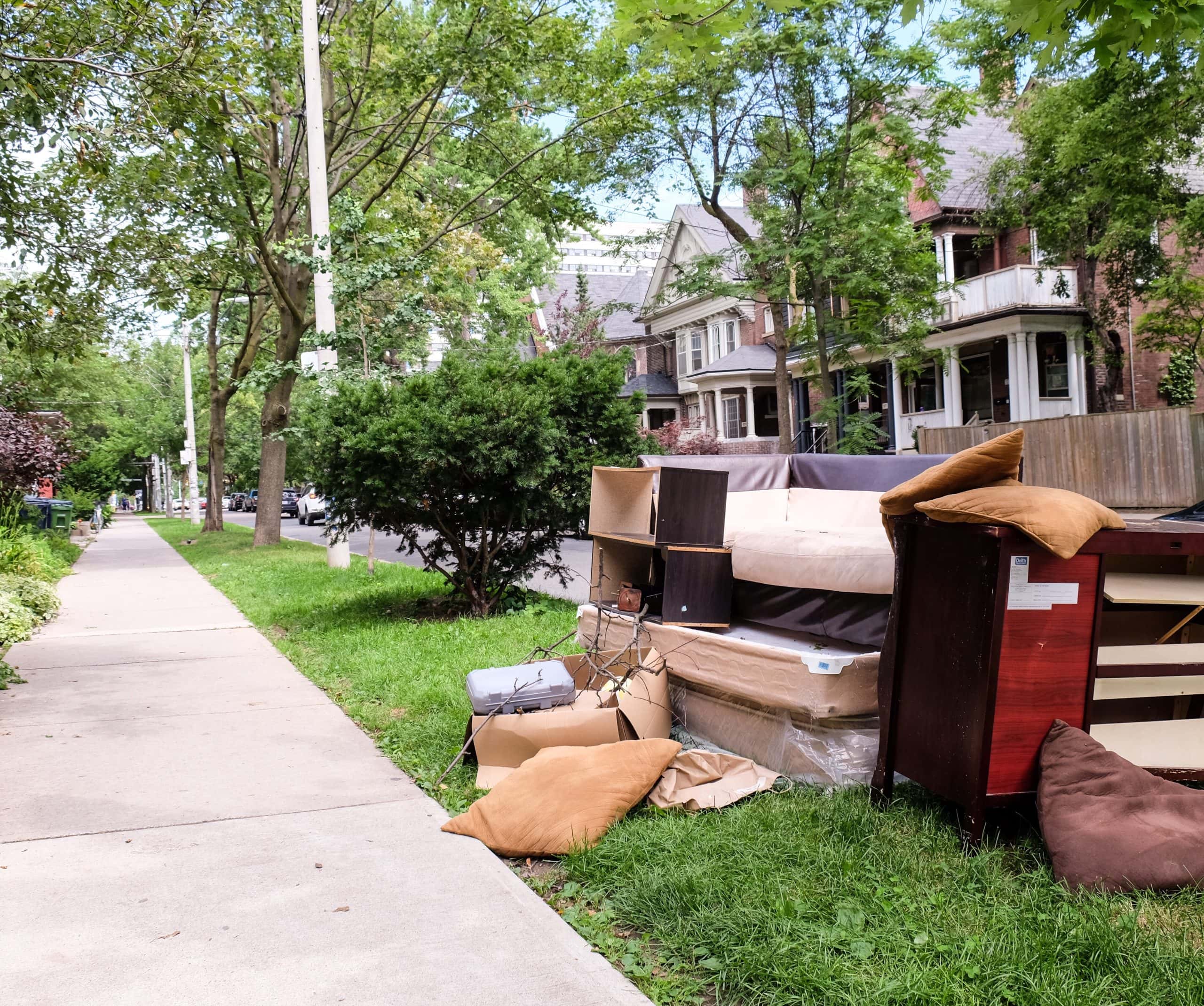 Old furniture, mattresses, and household items piled beside the sidewalk in front of residential homes, showing clutter and poor curb appeal in a neighbourhood street.