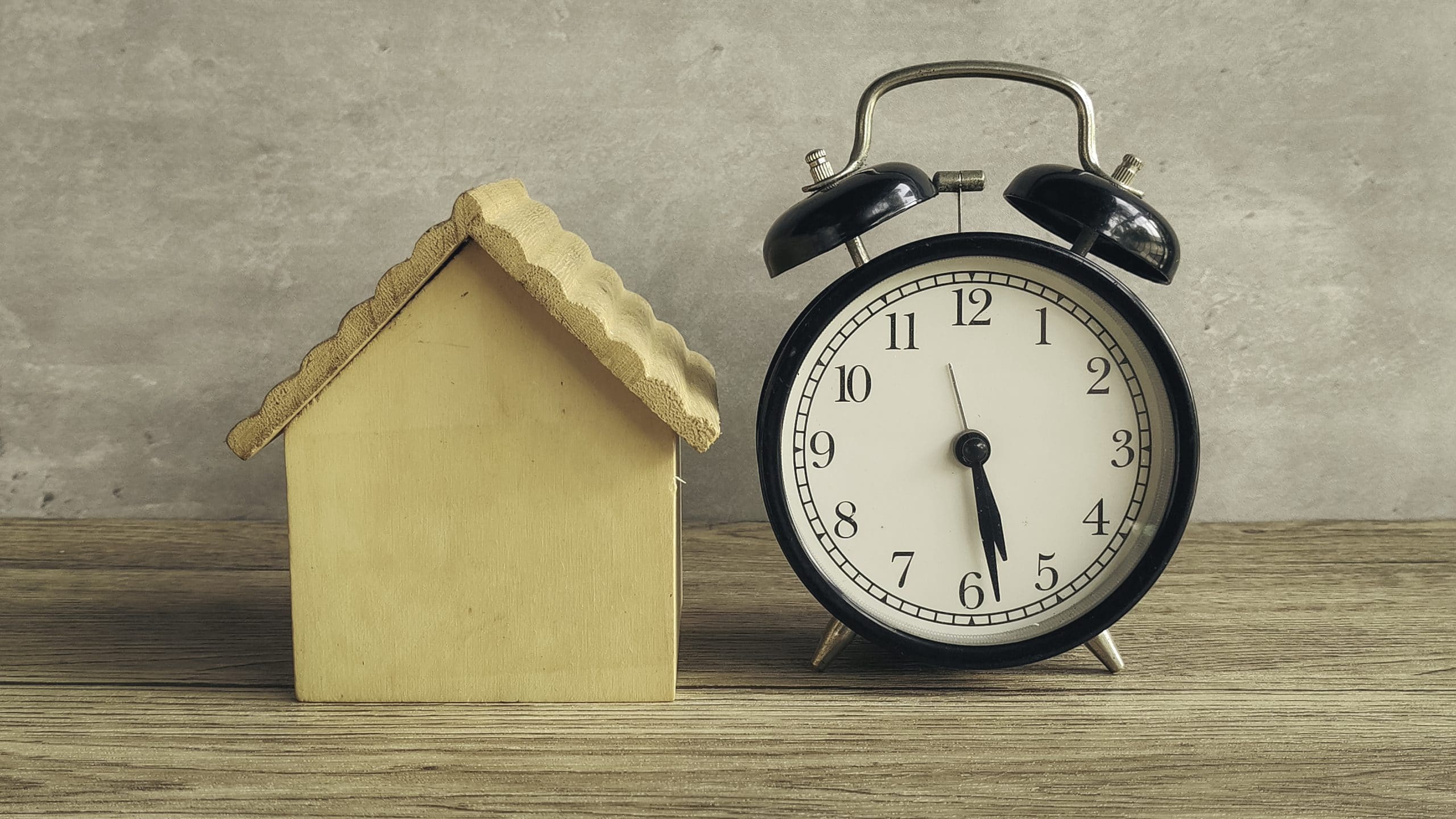 Small wooden house model beside a vintage alarm clock symbolizing real estate timing, mortgage deadlines, and the pressure of buying or selling a home.