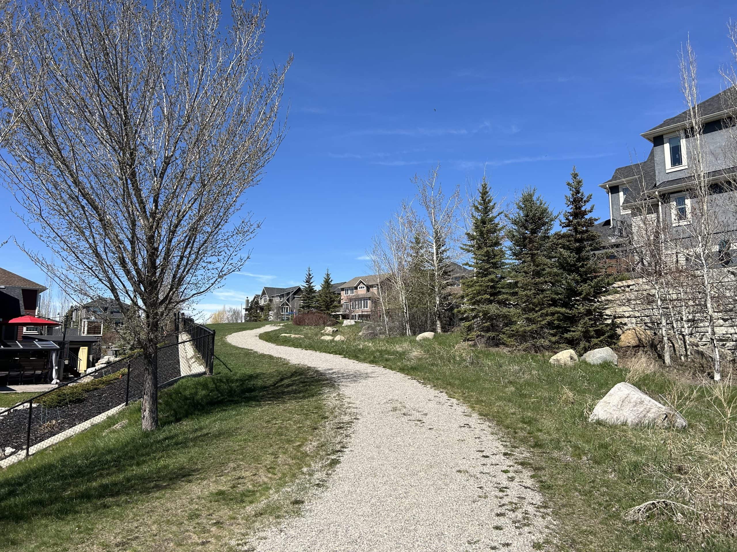 Walking path in Springbank Hill Calgary surrounded by upscale homes, mature trees, and landscaped green space, highlighting the quiet and scenic lifestyle of this west Calgary neighbourhood