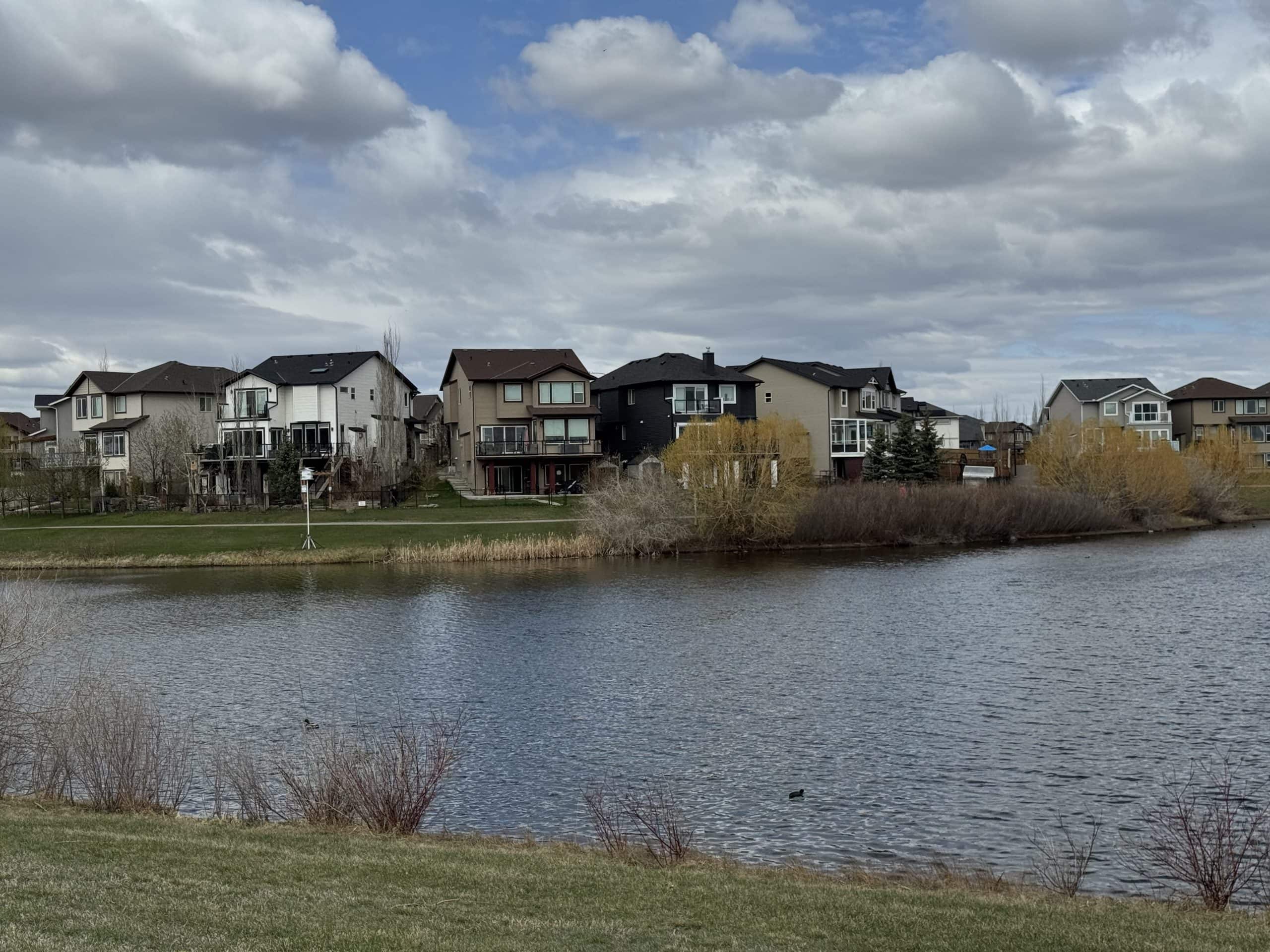 Luxury lakefront homes in a Calgary community under dramatic spring skies beside a peaceful stormwater pond and walking paths.