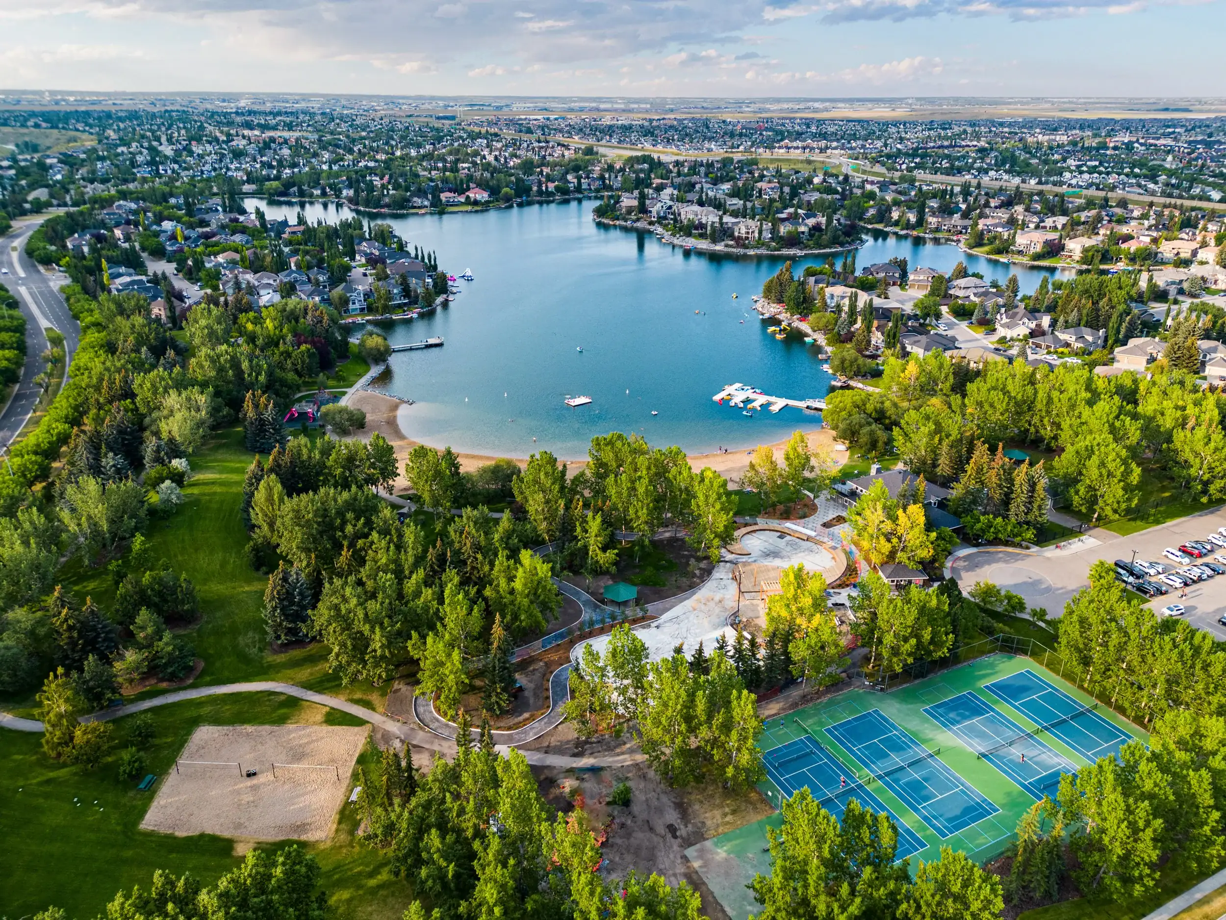 Aerial view of McKenzie Lake showcasing the private residents only lake, sandy beach, tennis courts, mature trees, and luxury waterfront homes that make this one of Calgary's most desirable family communities.