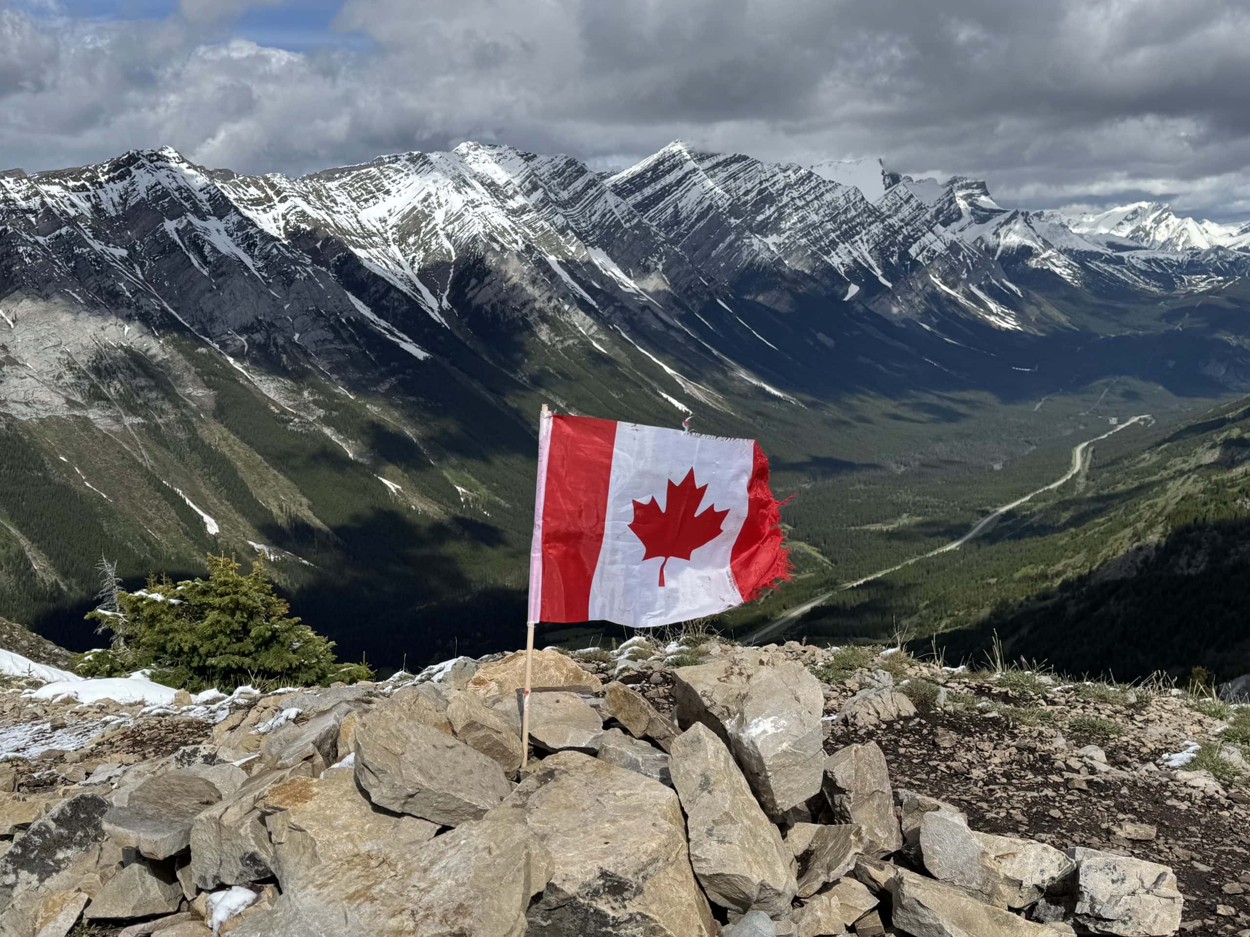 King Creek Ridge summit in Kananaskis with a Canadian flag overlooking the rugged Rocky Mountains and Highway 40.