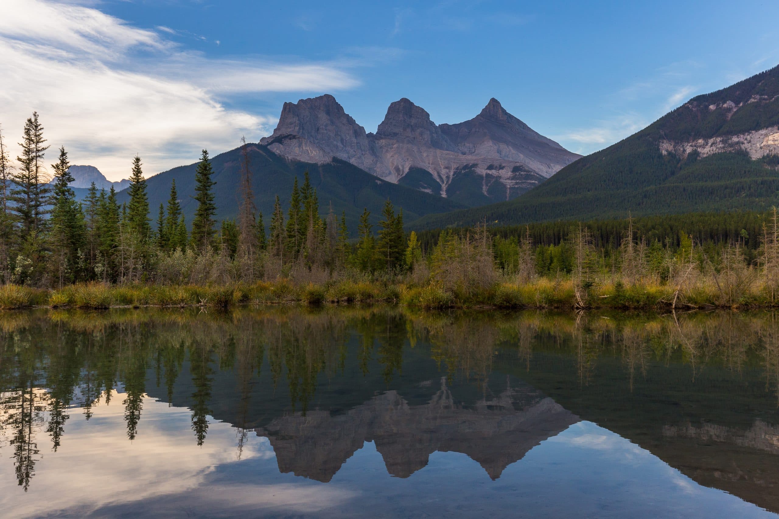 The iconic Three Sisters mountains reflected in calm water near Canmore, Alberta, surrounded by forest in the Canadian Rockies.