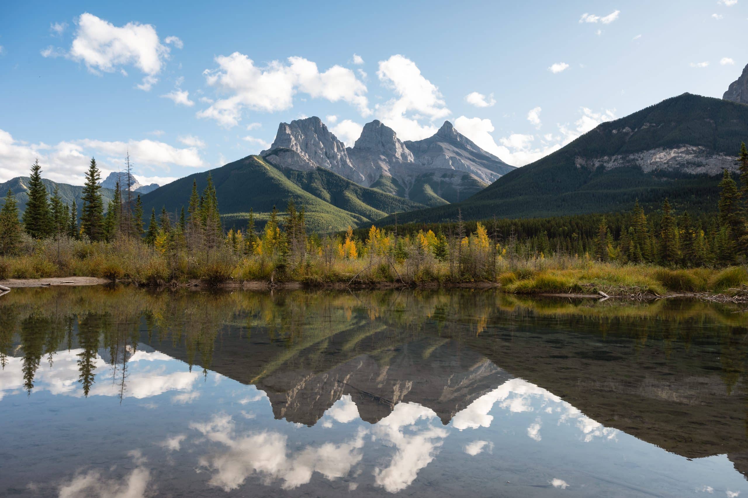 The Three Sisters mountains reflected in the water near Canmore, Alberta, surrounded by forest and fall colours in the Canadian Rockies.